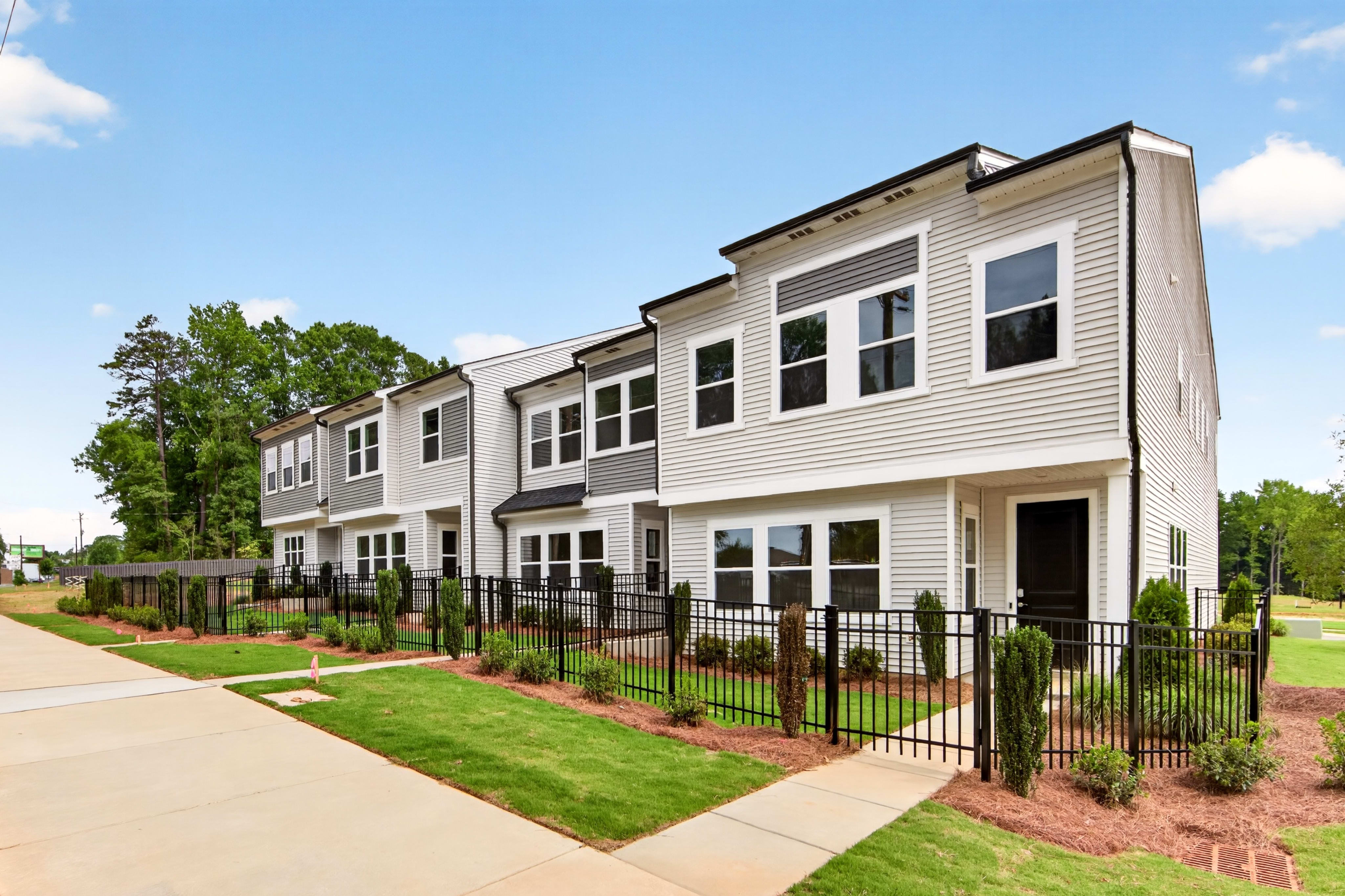 A row of modern townhouses with manicured lawns and black metal fences sits against a backdrop of trees and a clear blue sky.