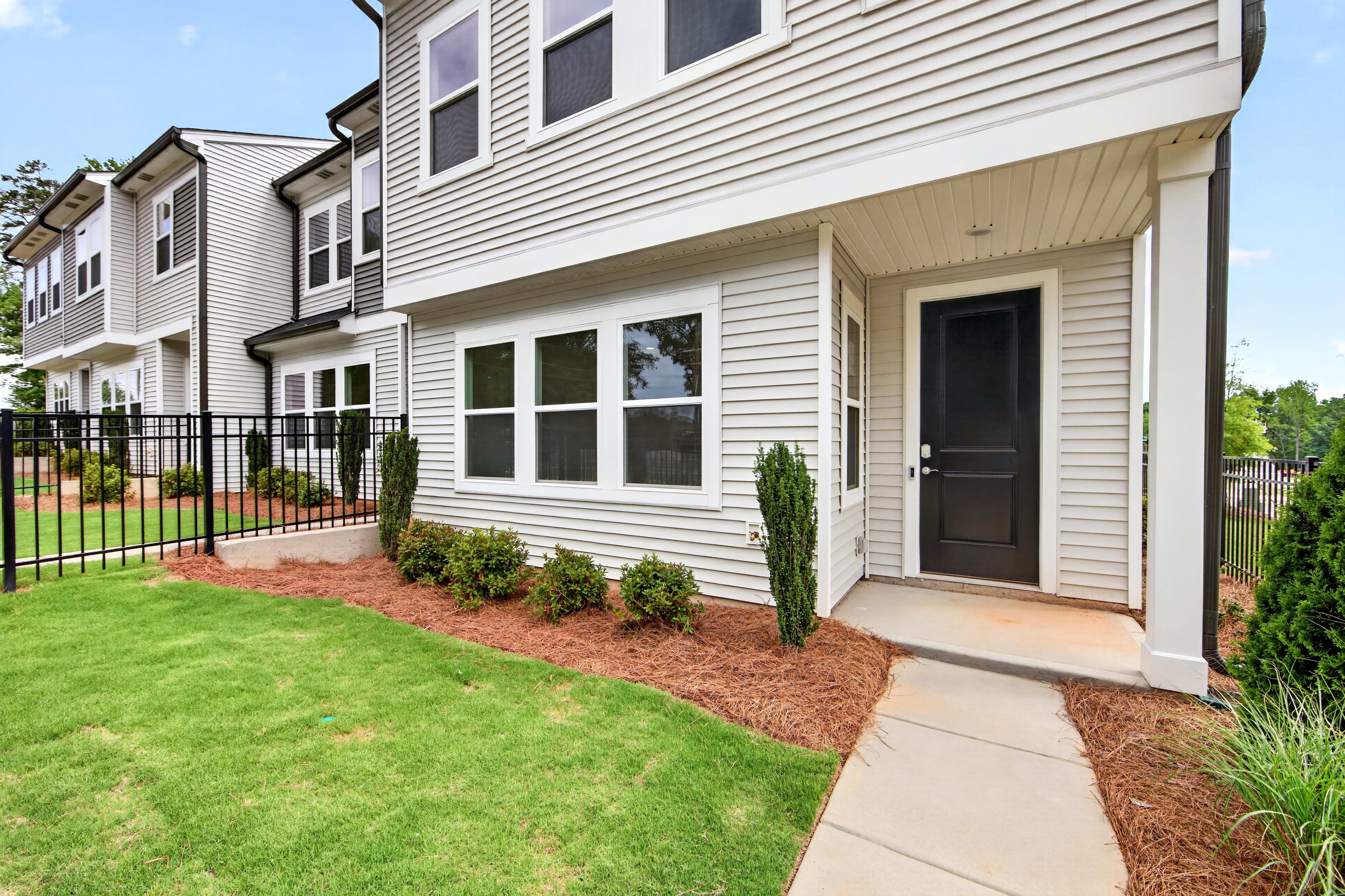 A neat lawn and walkway lead to the front door of a light gray townhouse, with more townhouses and trees visible in the background.