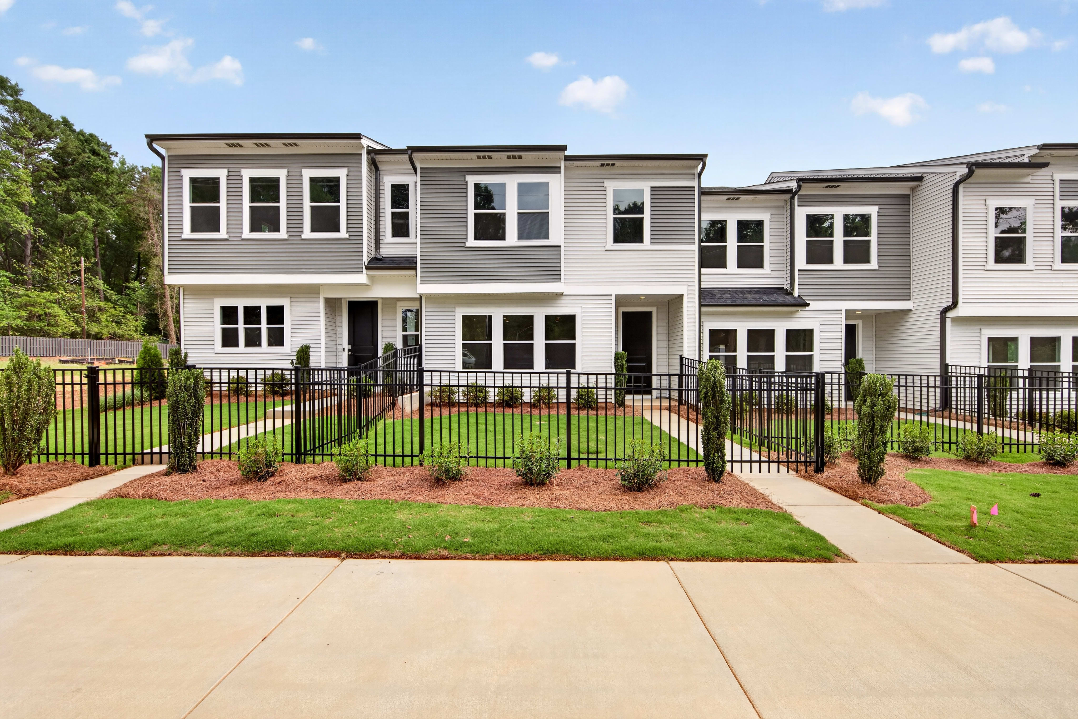 A row of modern townhouses with manicured lawns and a black metal fence in the foreground is set against a backdrop of trees and a bright blue sky.