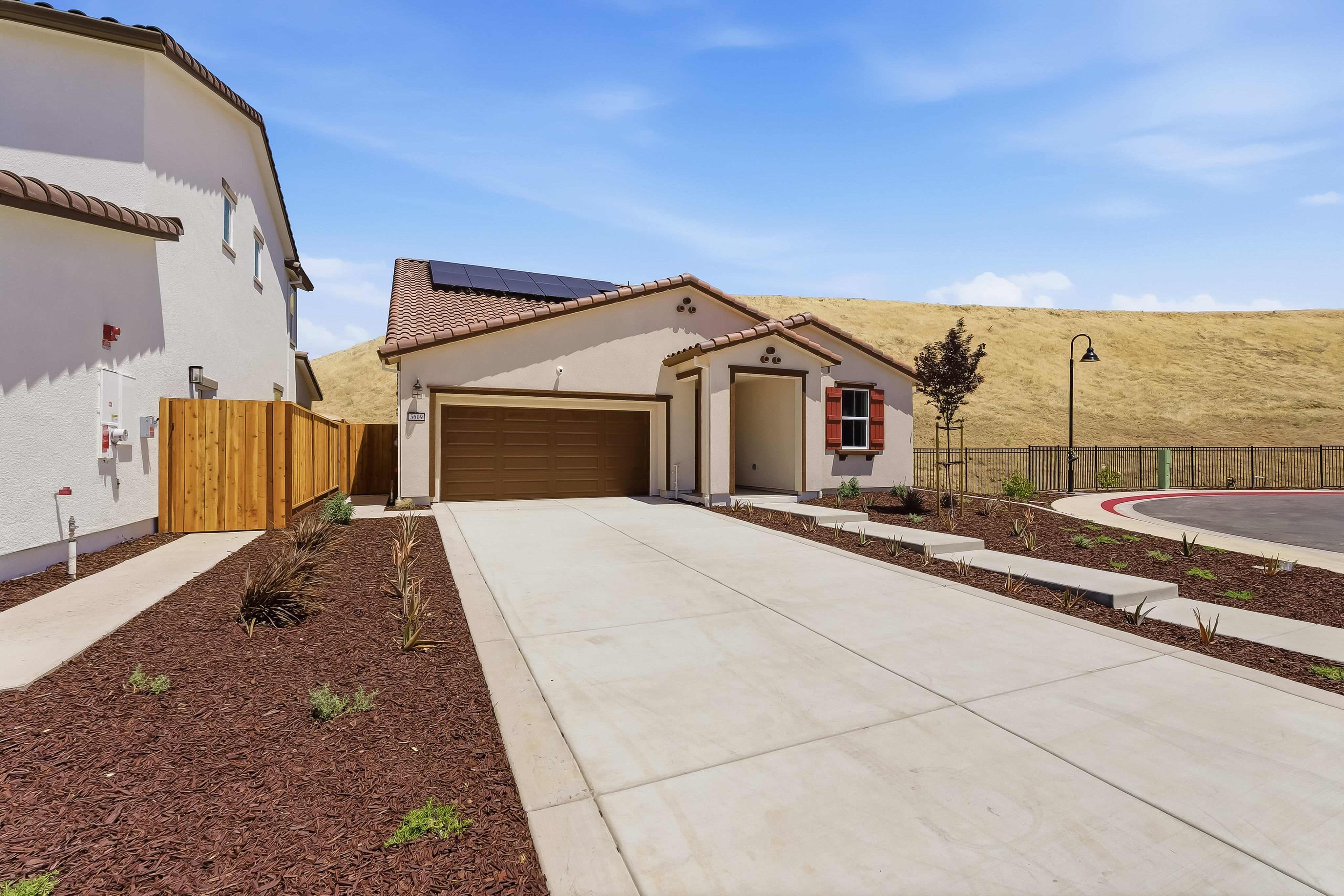 A modern beige house with solar panels sits on a concrete driveway, bordered by mulch and landscaping, with a rolling, grassy hill and a bright blue sky in the background.
