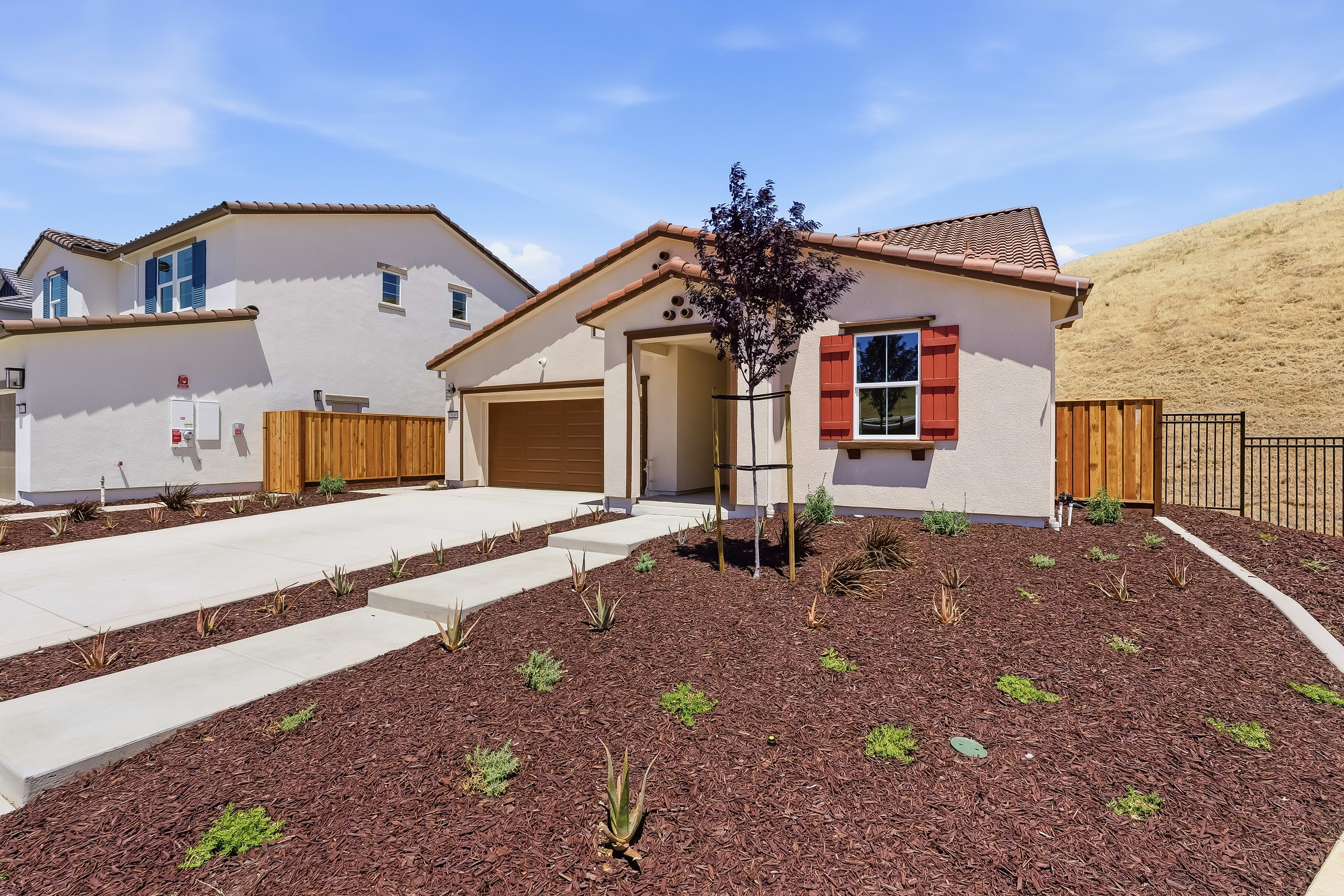 A stucco house with red shutters and a brown garage door sits on a landscaped lot with mulch and succulents in the foreground, while a grassy hill rises behind it under a blue sky.