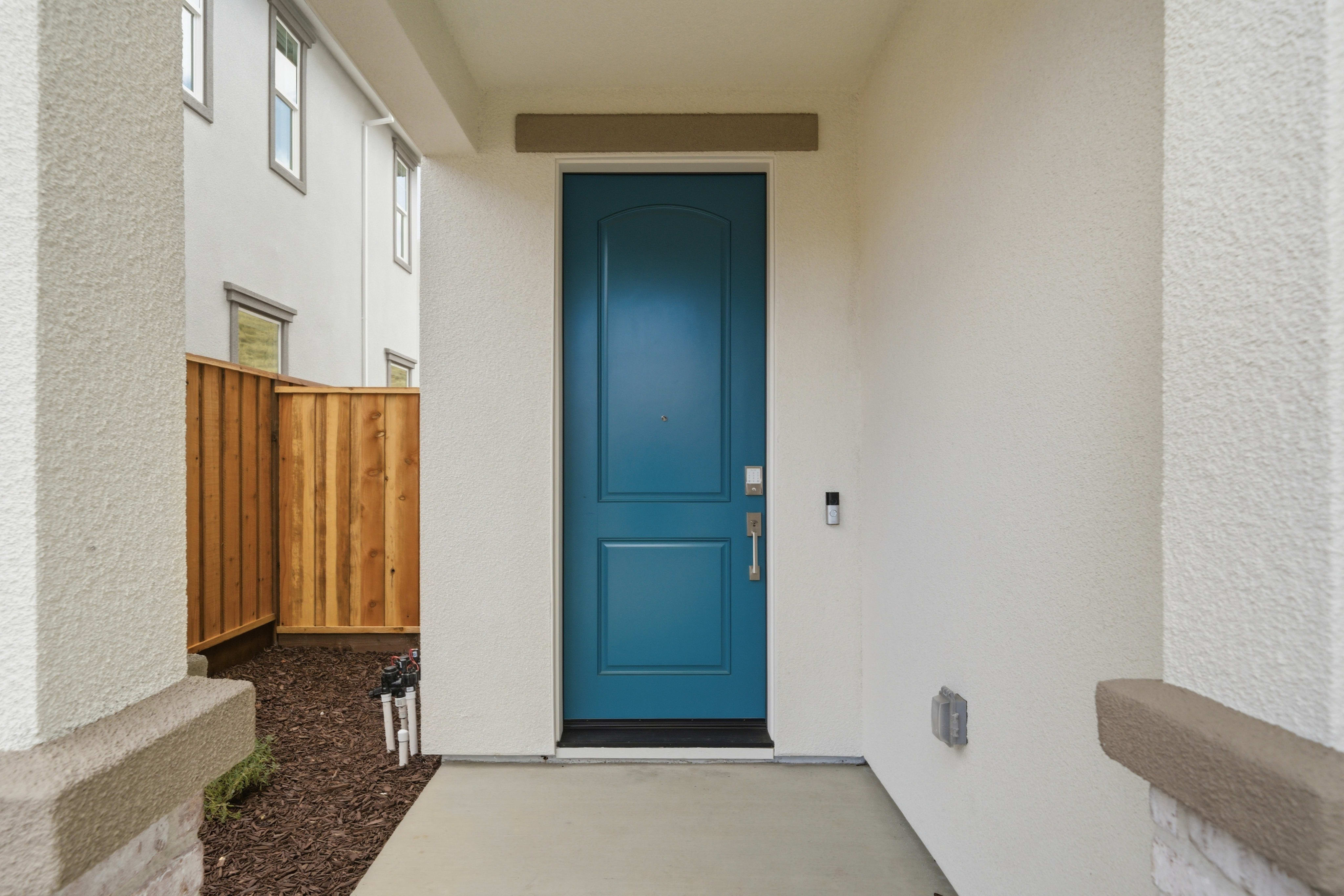 A teal front door stands on a white stucco house, with a wooden fence and neighboring houses visible in the background.