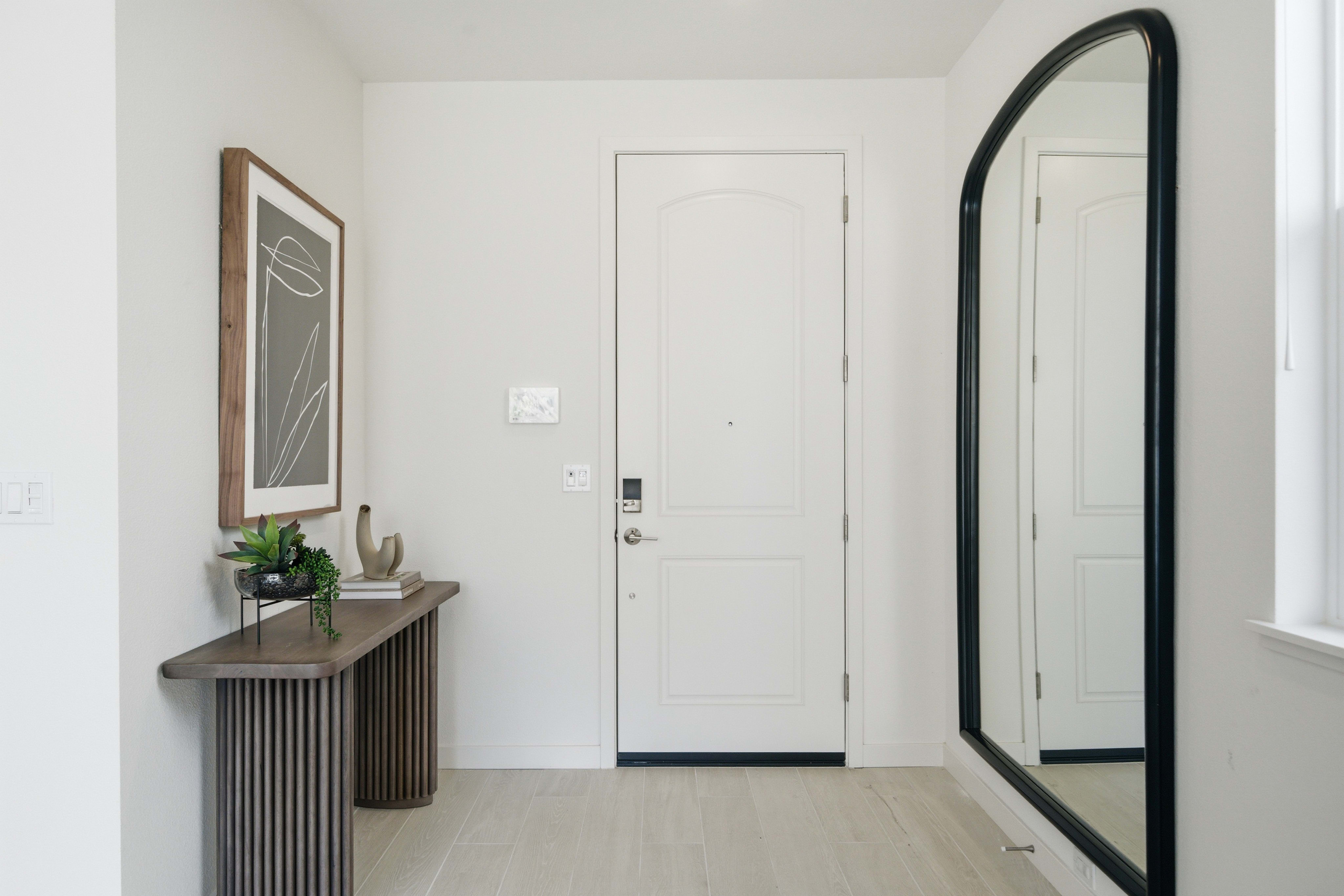 A modern entryway features a console table with decor in the foreground and a white door and large arched mirror in the background.