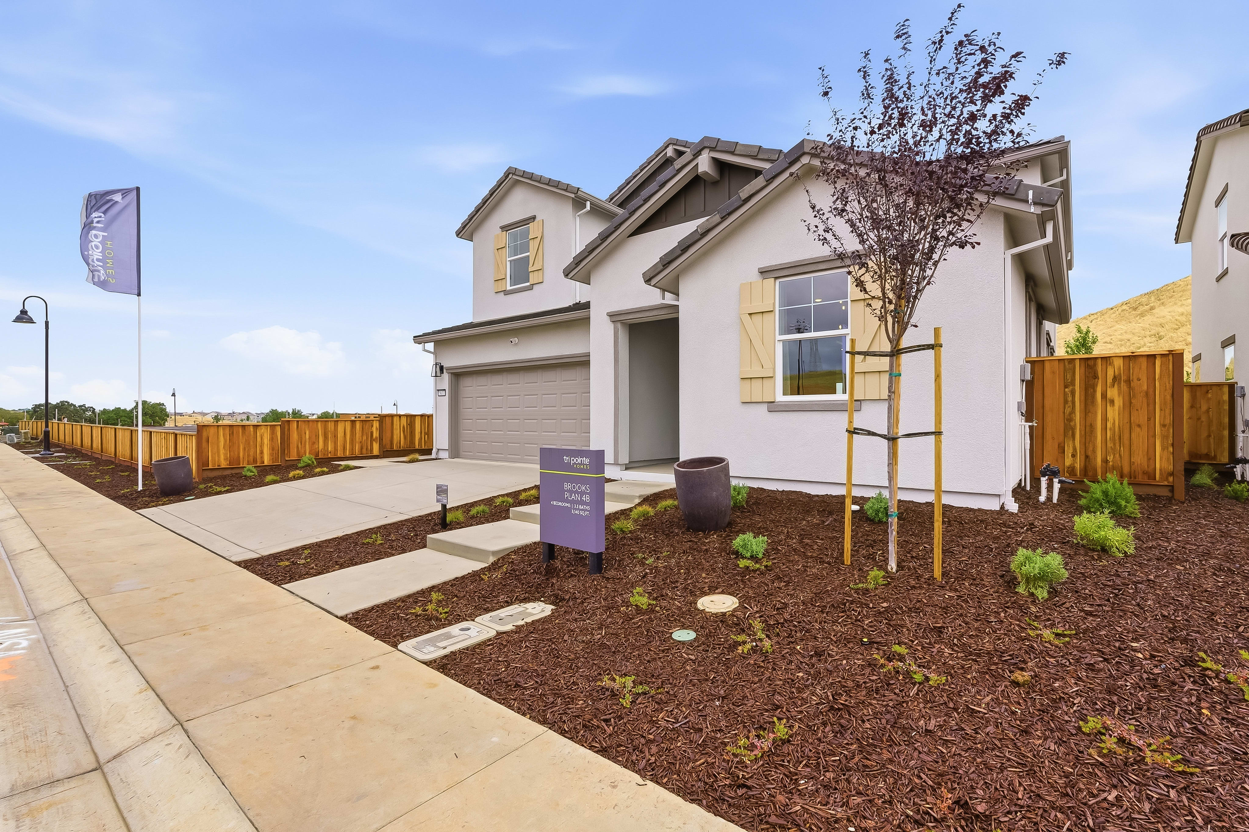 A modern, two-story house with light gray siding and tan shutters sits on a landscaped lot with a wooden fence, while a clear blue sky with a few clouds forms the background.