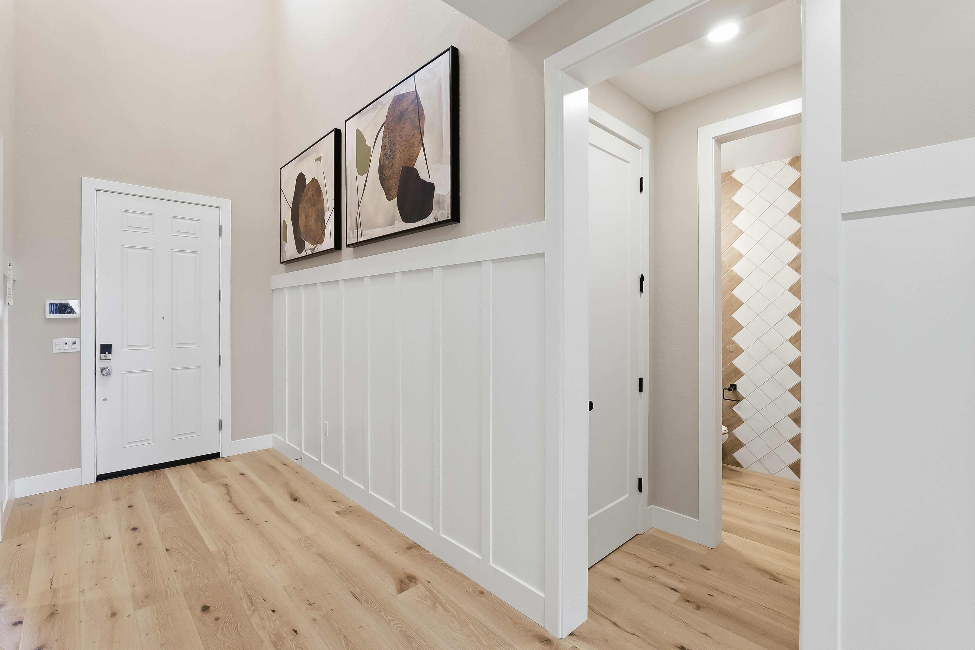 A bright hallway with light wood floors and white wainscoting in the foreground leads to a doorway revealing a room with patterned tile walls in the background.