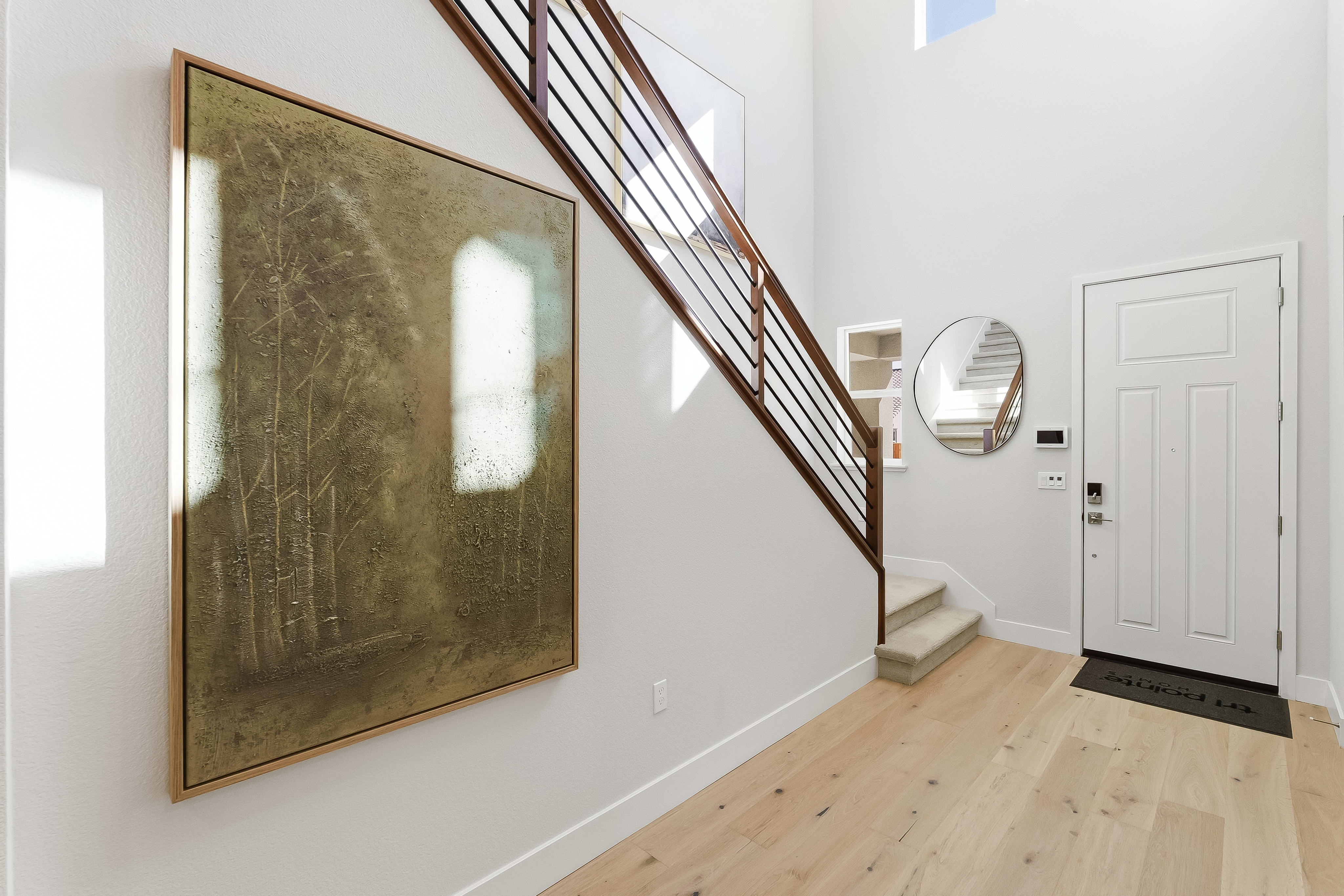 A large abstract painting hangs on the wall next to a staircase with a wooden railing, while a white door and a round mirror are visible in the background.