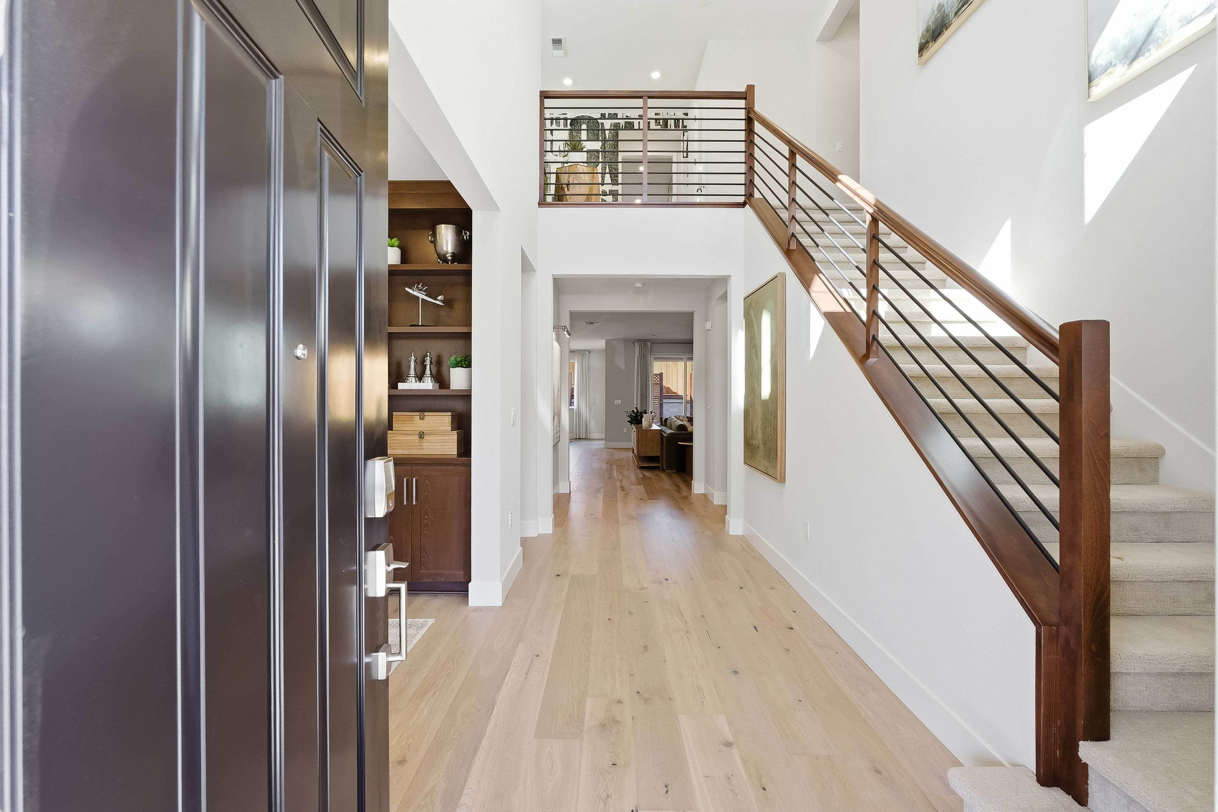 A dark wooden door opens into a bright, modern foyer with a light wood floor, a staircase with a metal railing, and a hallway leading to the back of the house.