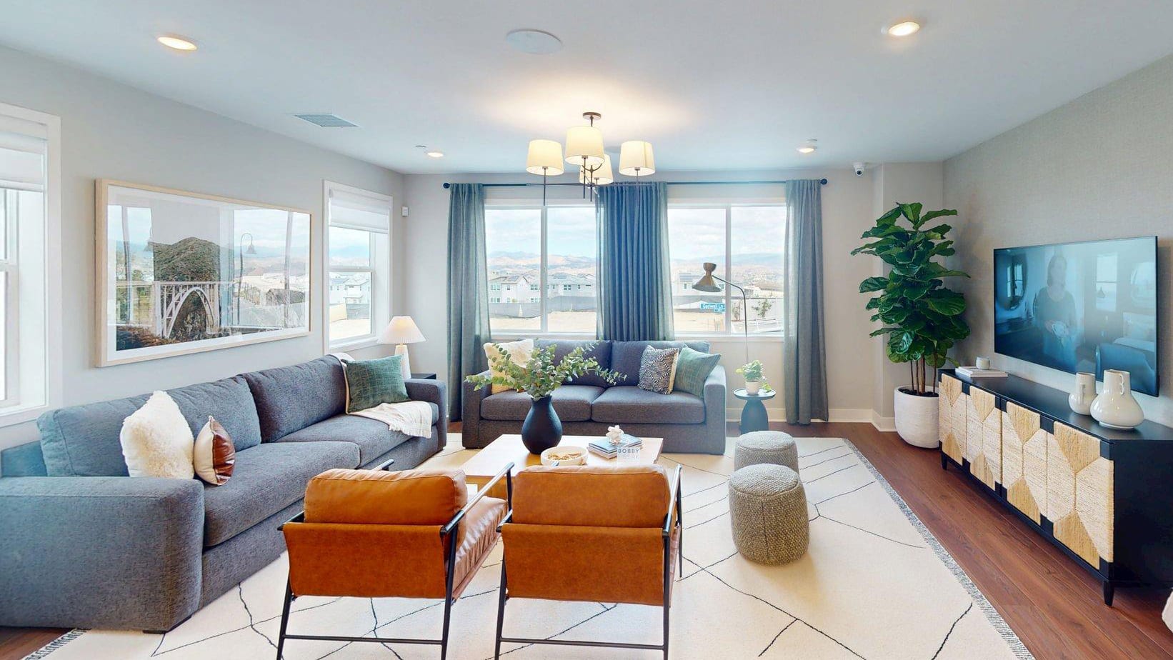 A spacious living room with a large gray sectional sofa and two brown leather armchairs in the foreground, and large windows with a view of a distant landscape in the background.