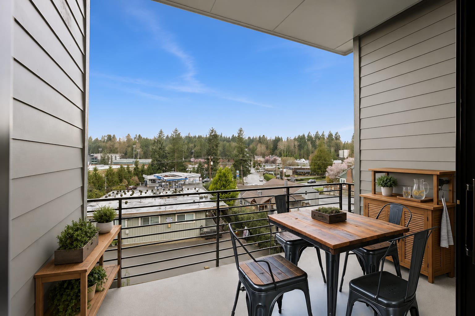 A balcony with a dining set in the foreground overlooks a suburban landscape with trees and buildings under a bright blue sky.