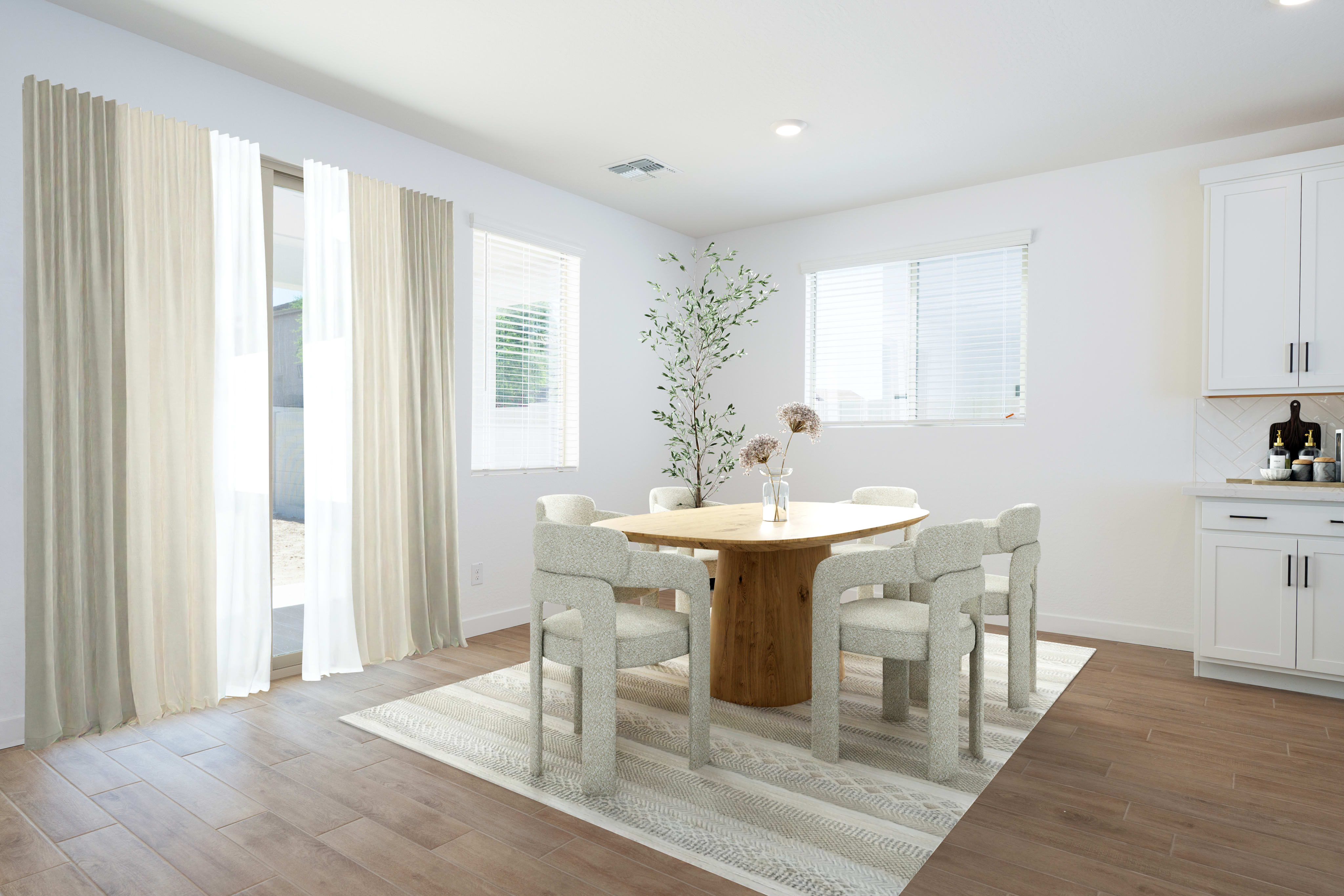 A light wood dining table with chairs sits on a striped rug in the foreground, while a bright room with windows and white cabinets is visible in the background.