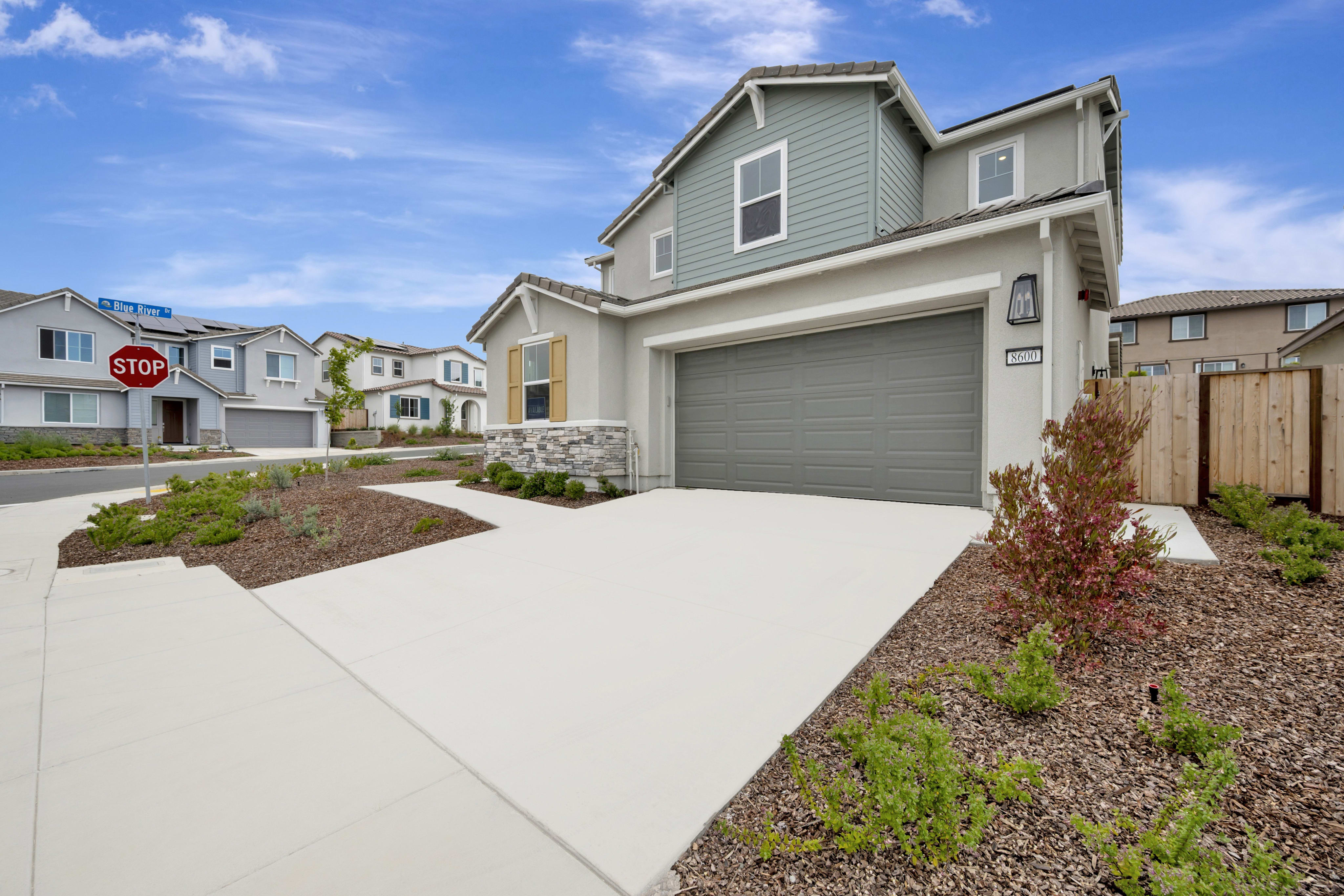 A modern two-story house with a gray garage door and landscaping in the foreground sits under a blue sky with clouds, while other houses line the street in the background.