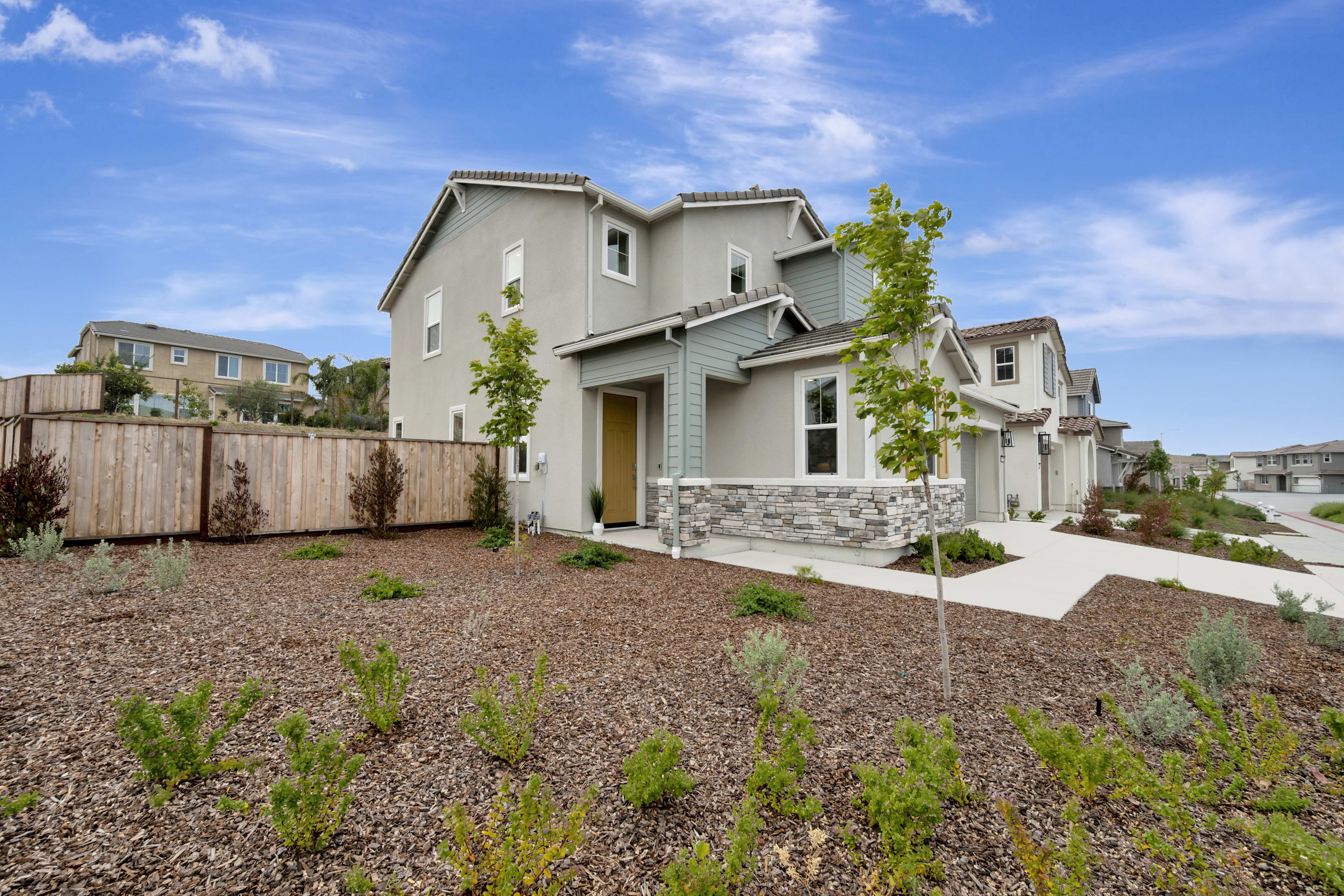 A modern, two-story house with a stone-accented lower level stands behind a landscaped yard with mulch and low-lying plants, while other houses and a wooden fence line the background under a cloudy blue sky.