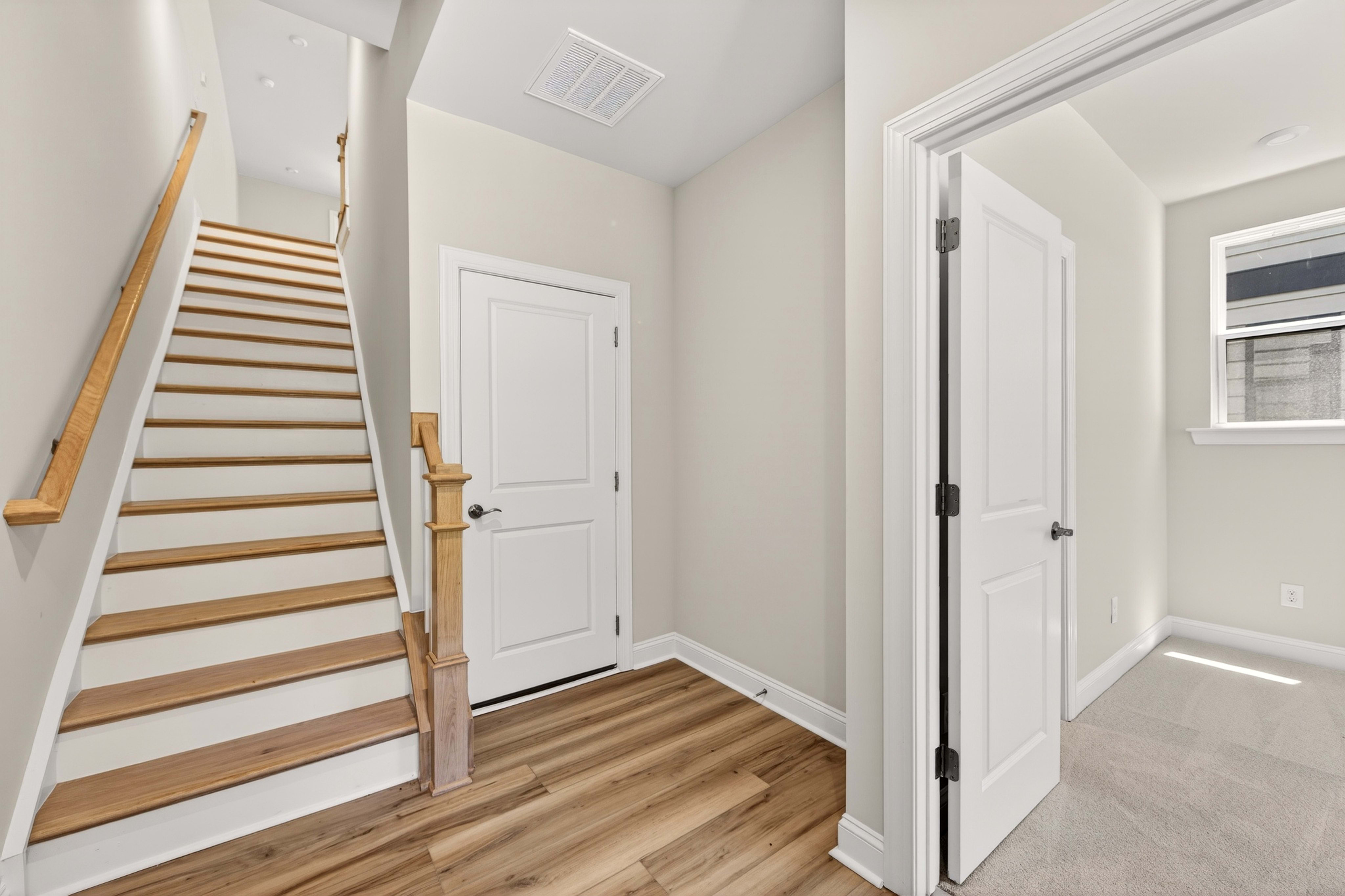 A wooden staircase and floor are in the foreground, leading to a hallway with white doors and a bright room in the background.