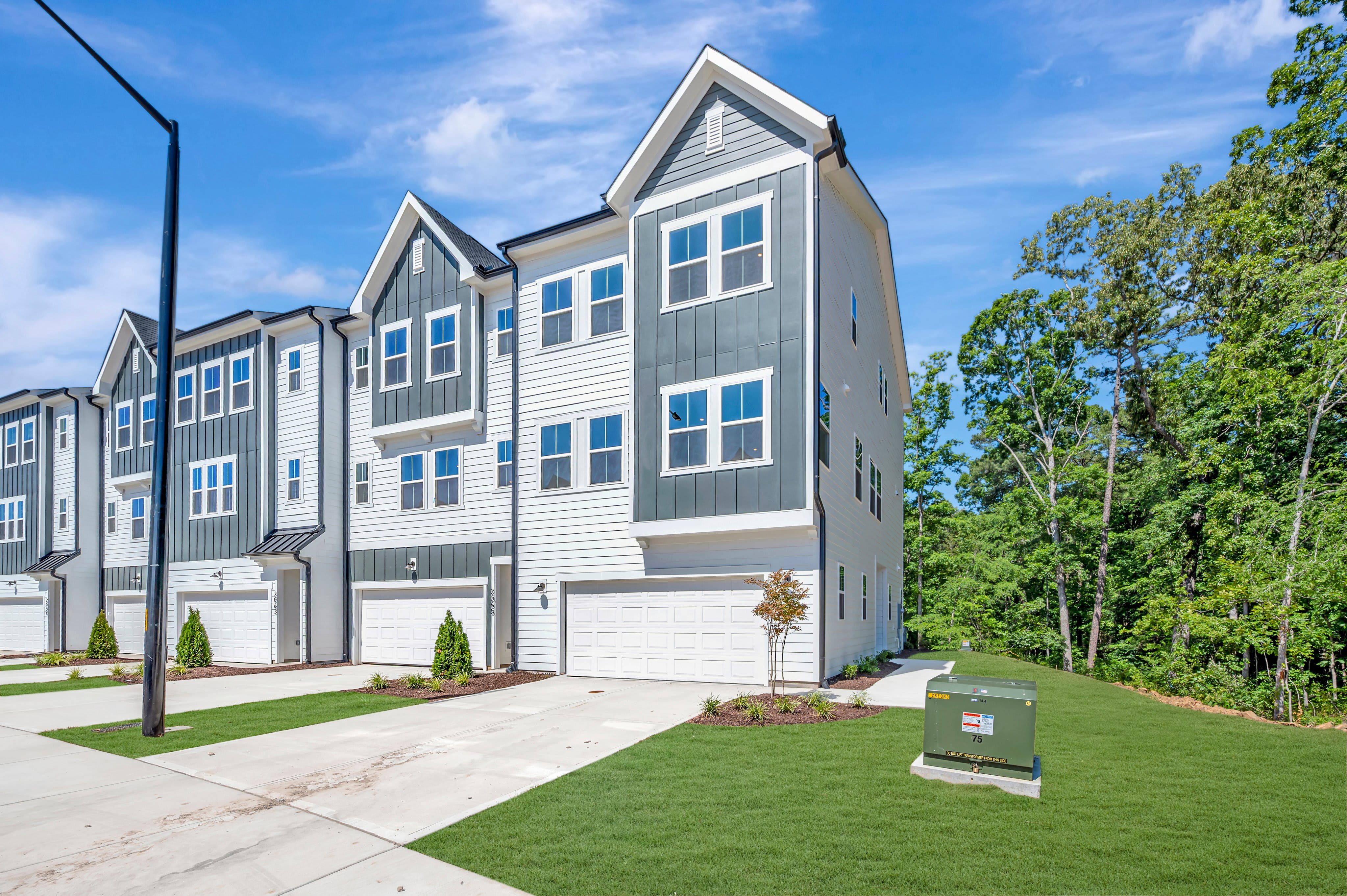 Modern townhouses with manicured lawns and a utility box in the foreground are set against a backdrop of lush green trees under a bright blue sky.