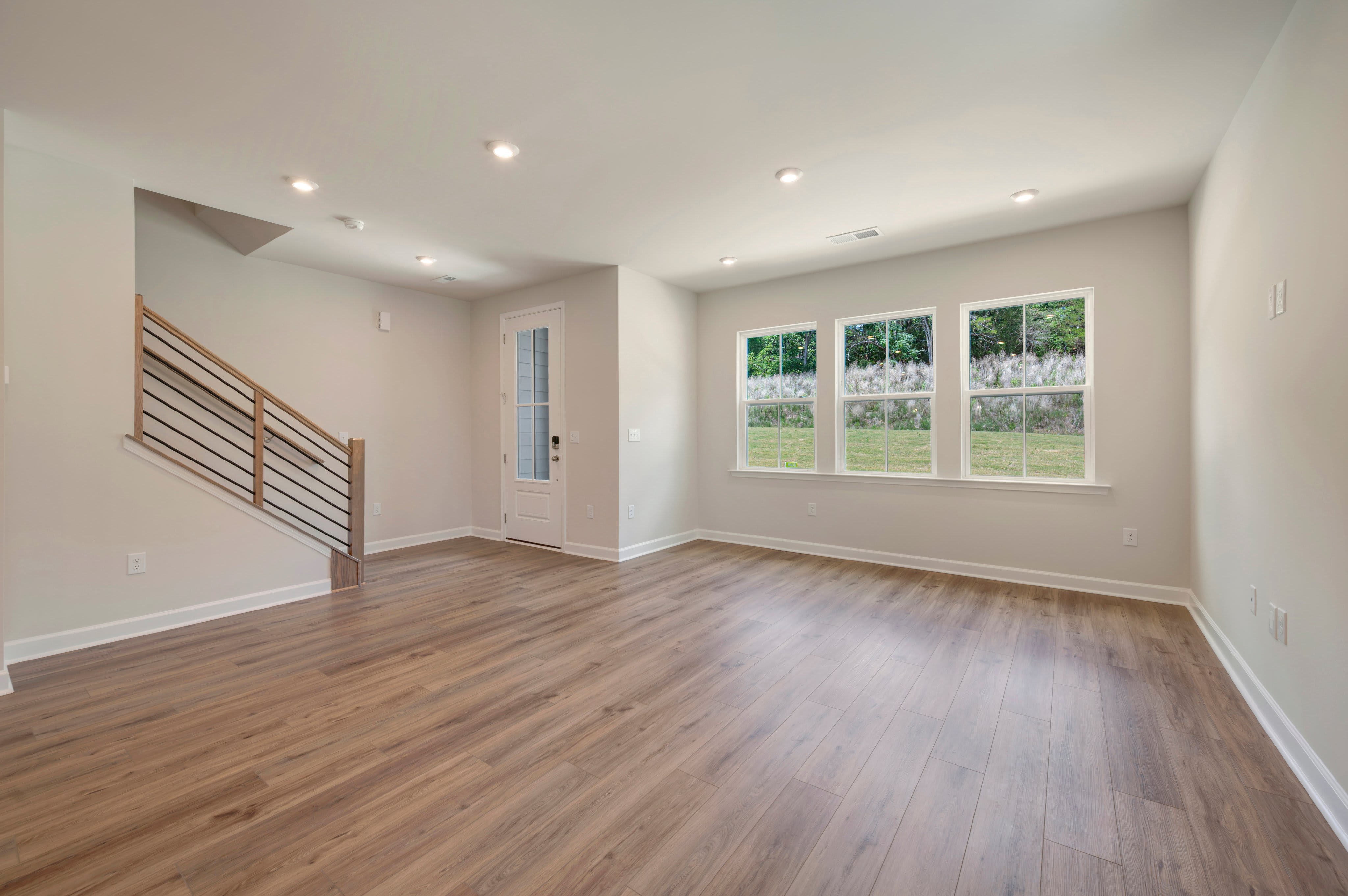 A modern living room with a wooden floor and a staircase in the foreground, and three windows overlooking a green landscape in the background.