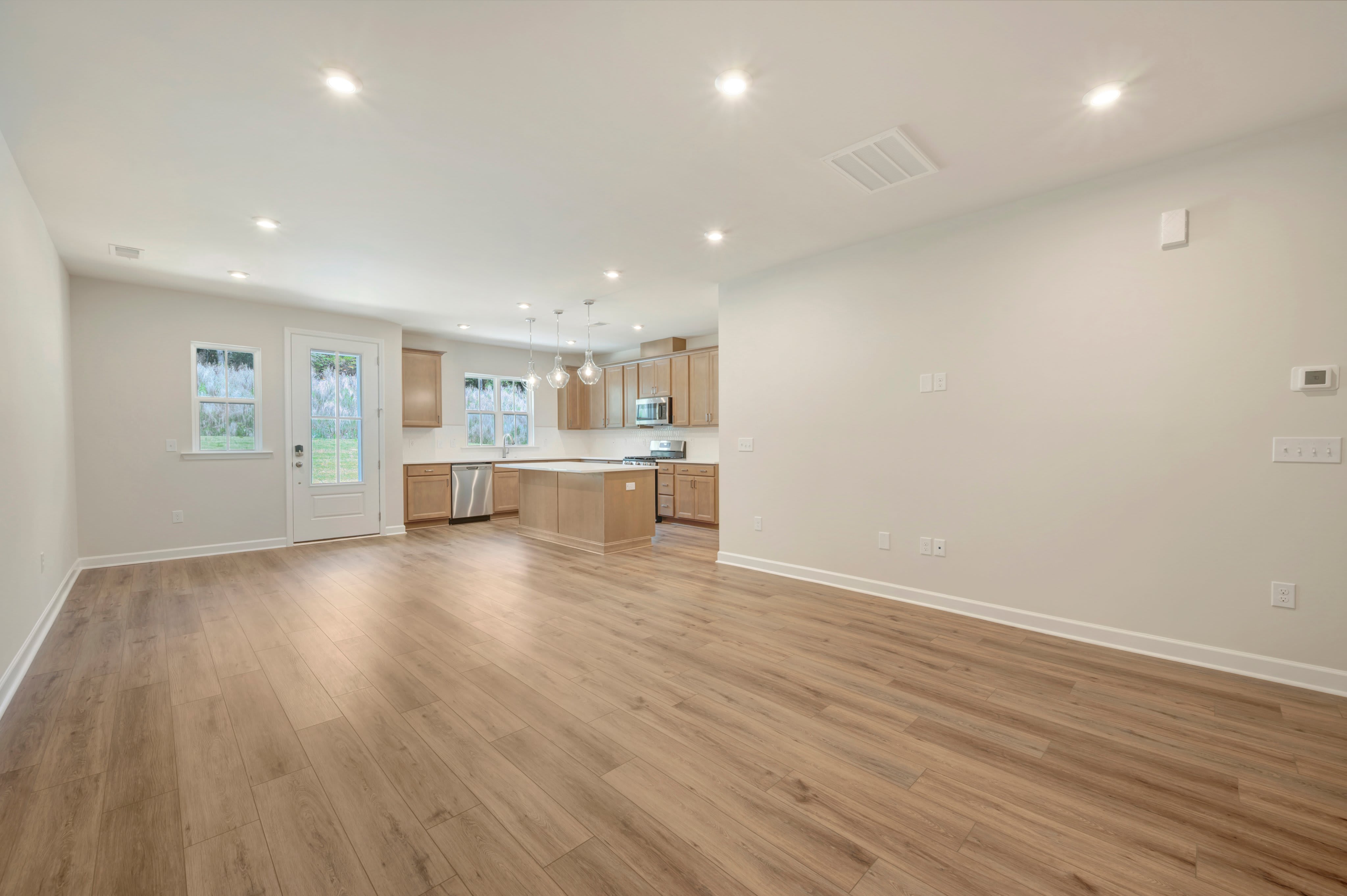 A spacious, modern living area with light wood flooring in the foreground opens to a well-lit kitchen with light wood cabinets and an island in the background.