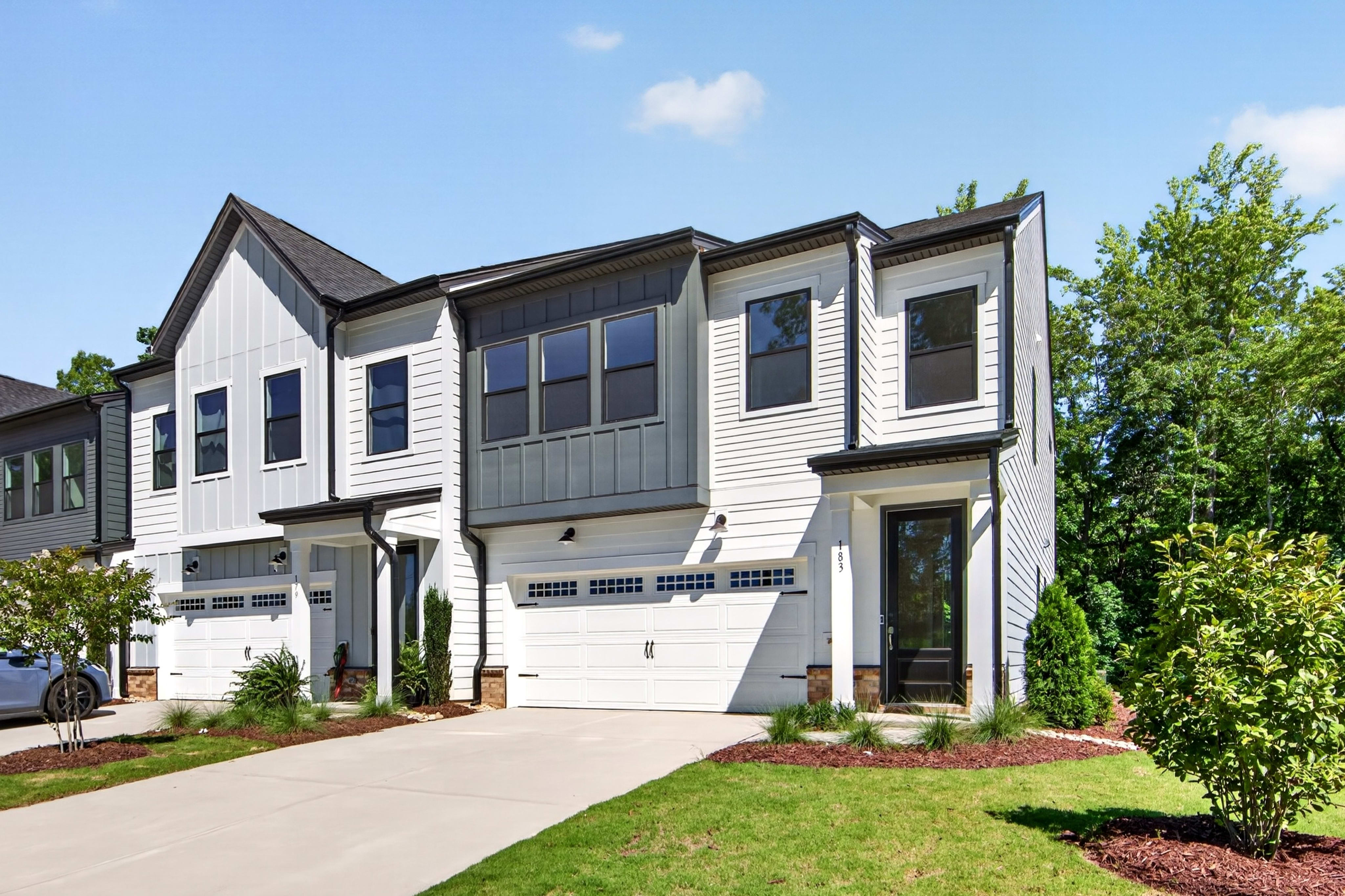 A modern townhouse with white siding and dark trim stands in the foreground, with lush green trees and a clear blue sky in the background.