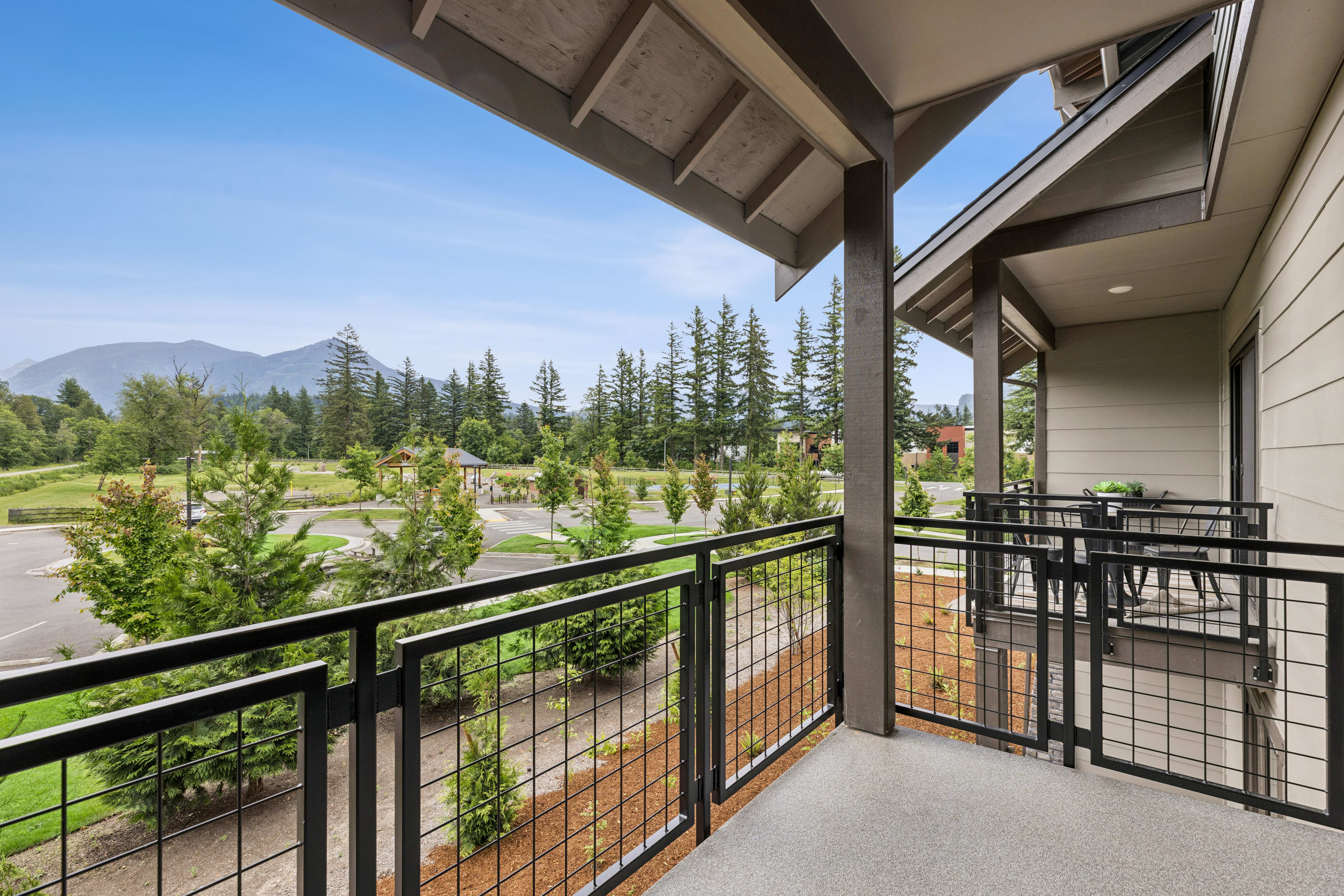 A modern balcony with a metal railing overlooks a scenic landscape of trees, mountains, and a clear blue sky.