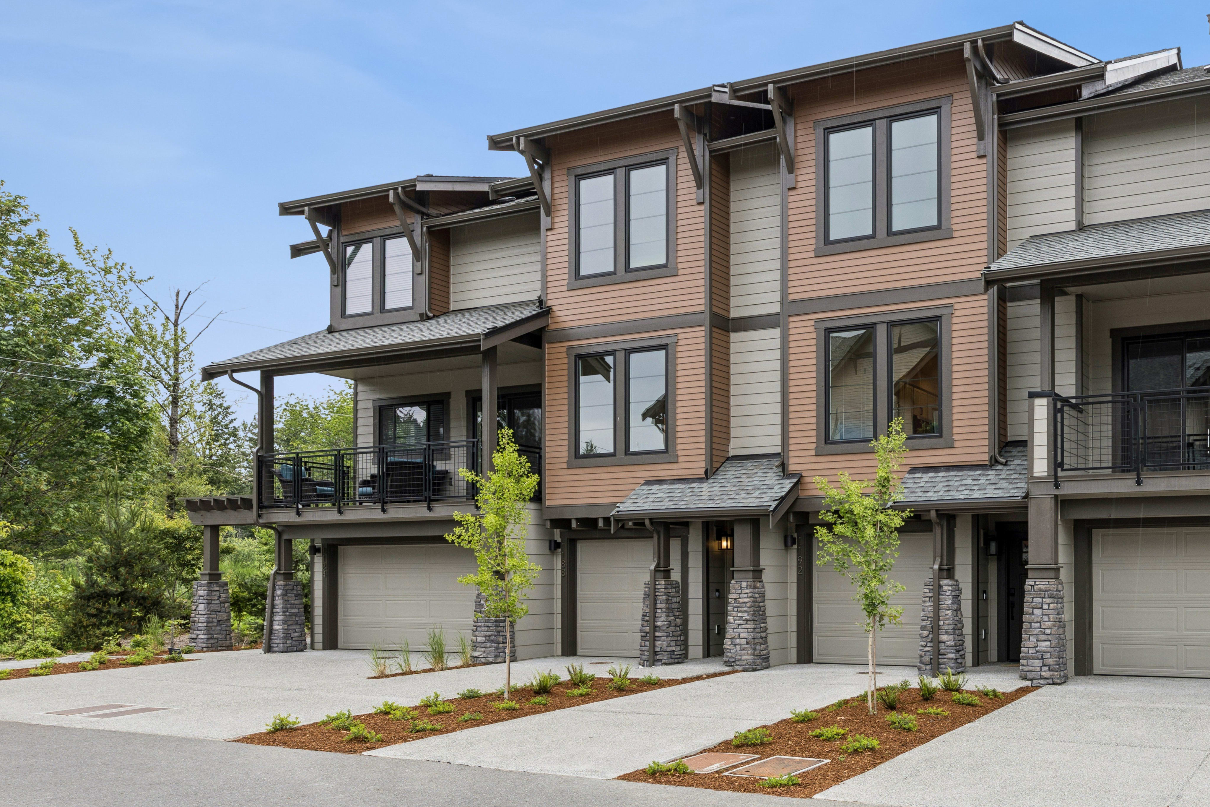 Modern townhouses with attached garages and manicured landscaping are set against a backdrop of lush green trees under a clear sky.