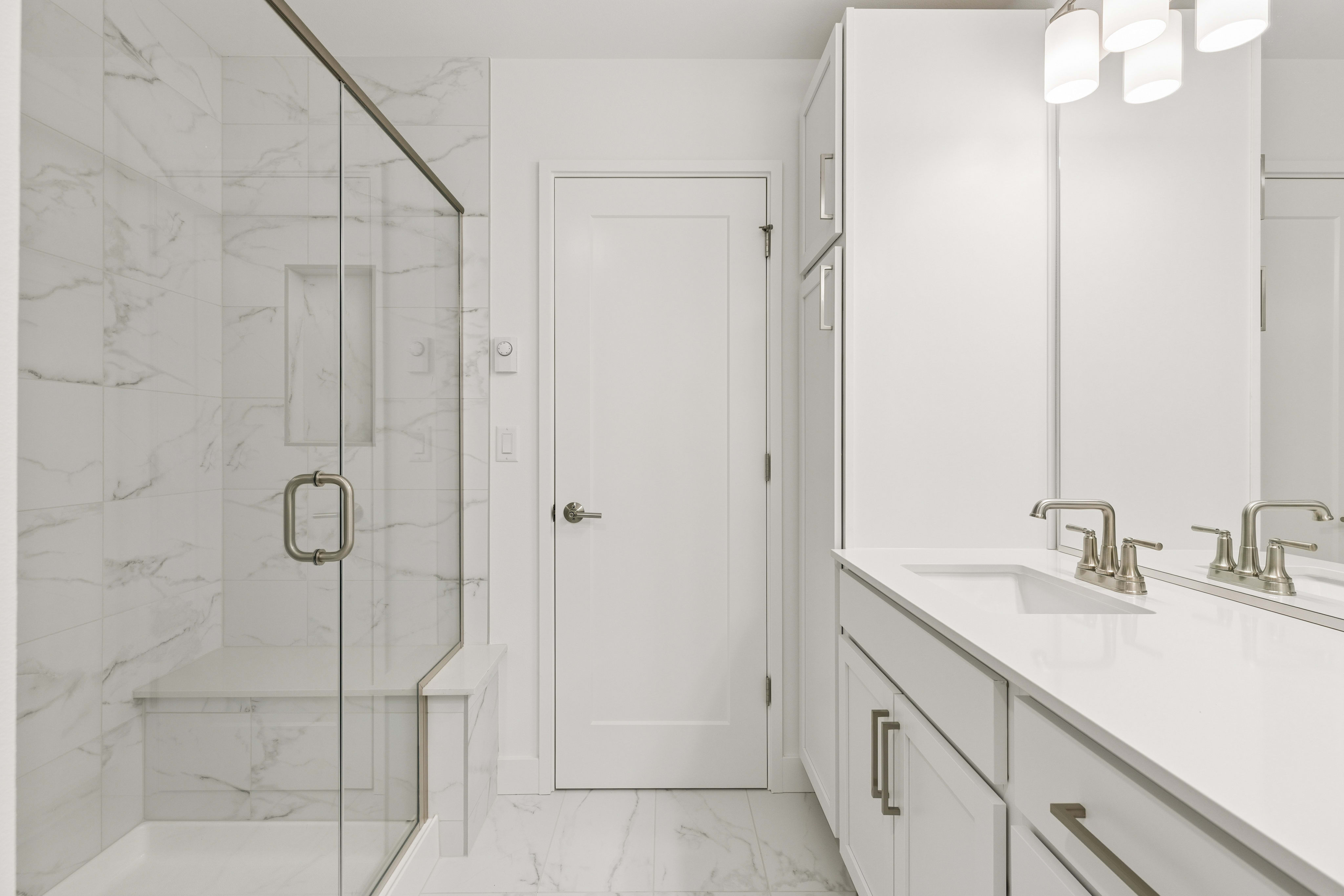 A modern white bathroom features a glass-enclosed shower with marble tile in the foreground and a double vanity with white countertops and cabinets in the background.