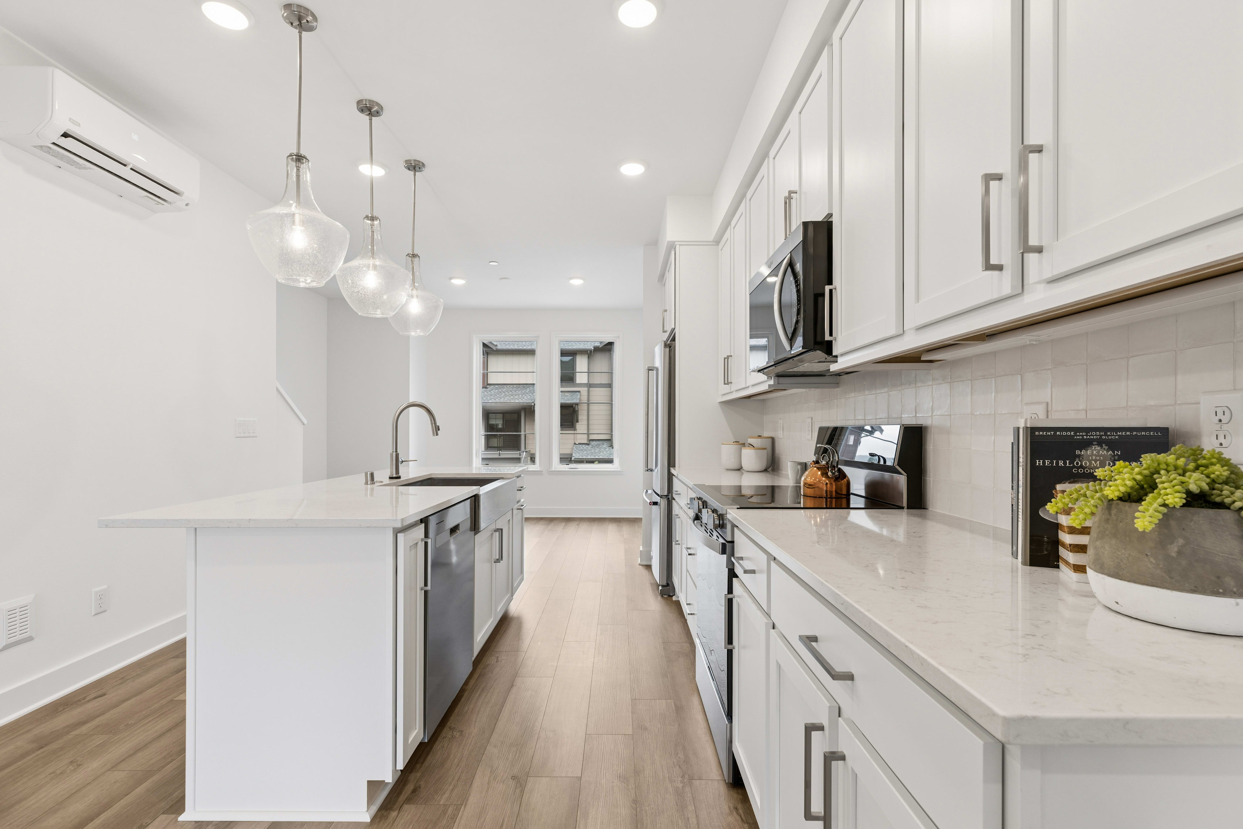 A modern white kitchen with a large island in the foreground and a hallway with windows in the background.