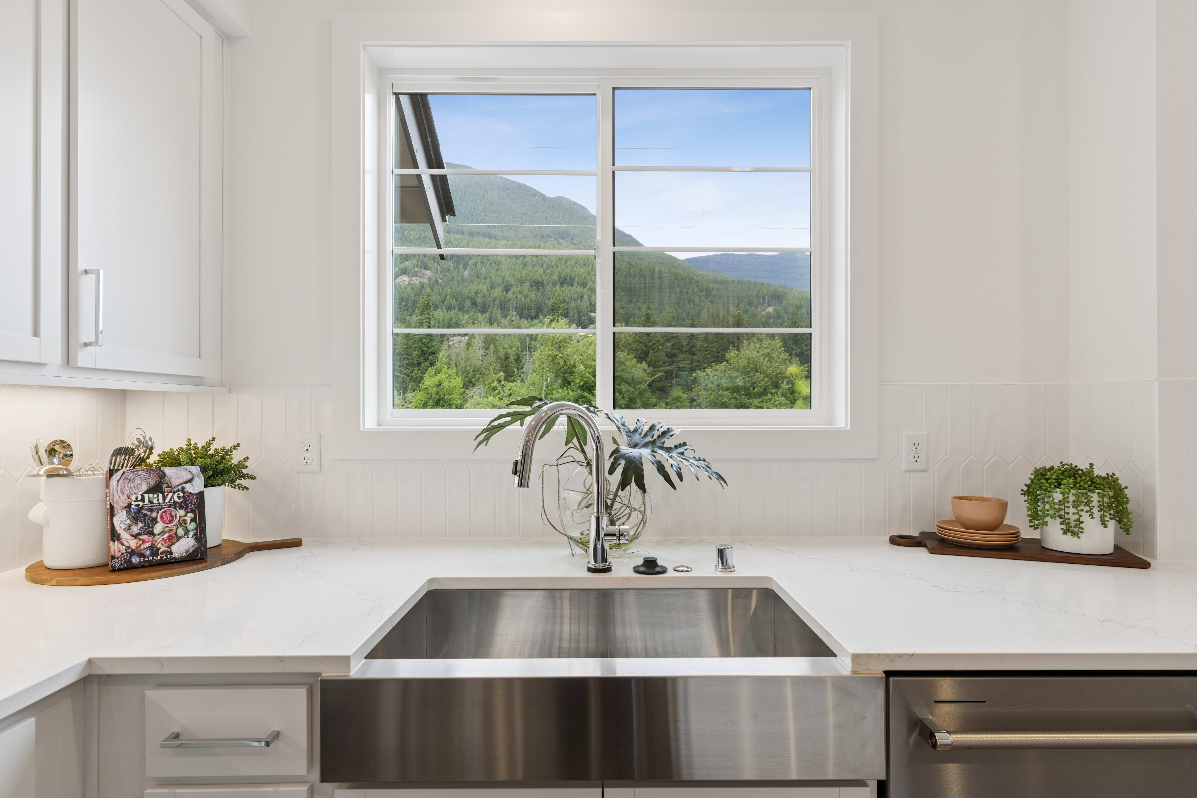 A modern kitchen sink with a stainless steel basin is in the foreground, while a lush green mountain landscape is visible through a large window in the background.