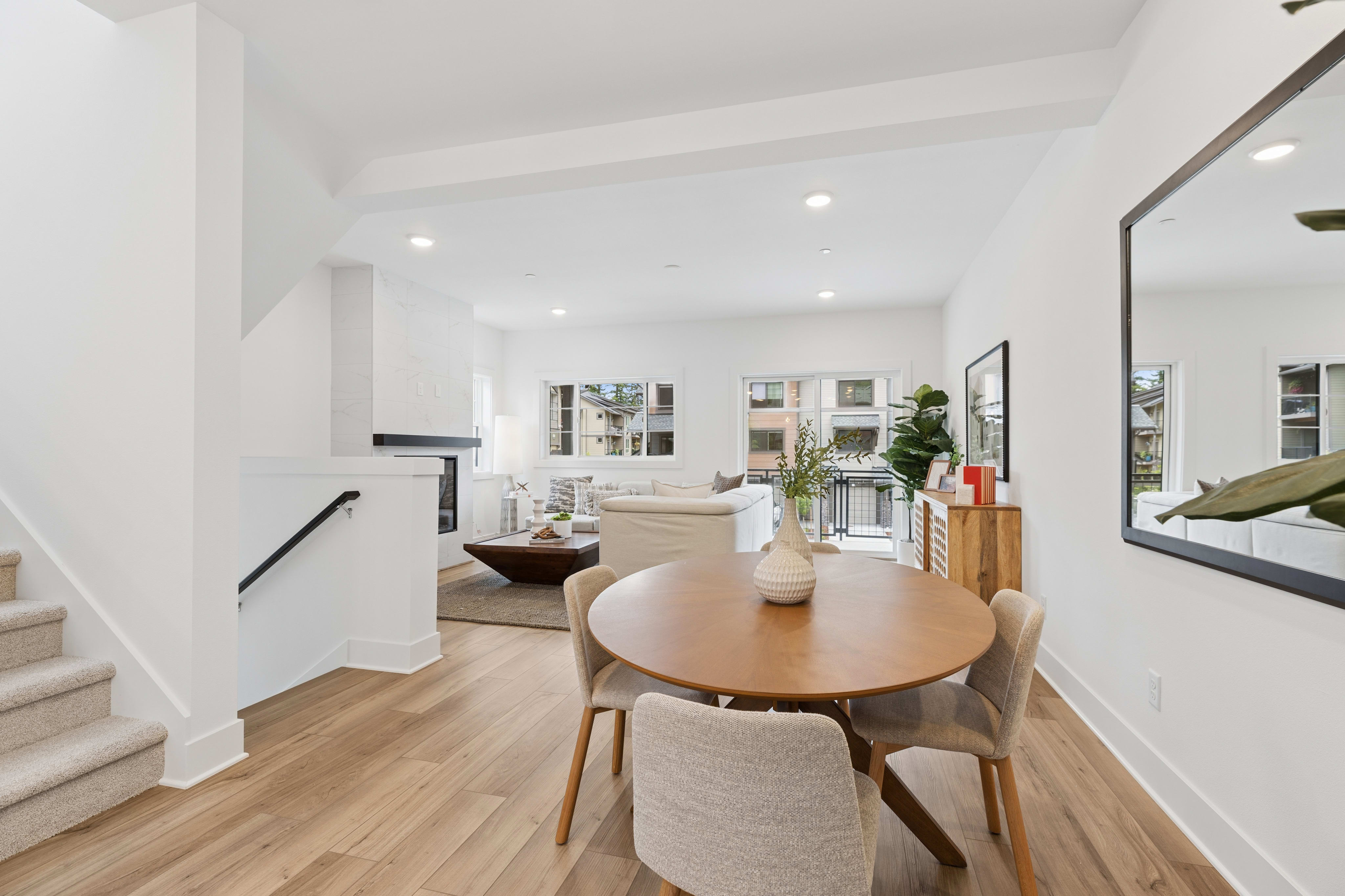 A modern dining area with a round table and chairs sits in the foreground, while a spacious living room with a fireplace and large windows is visible in the background.