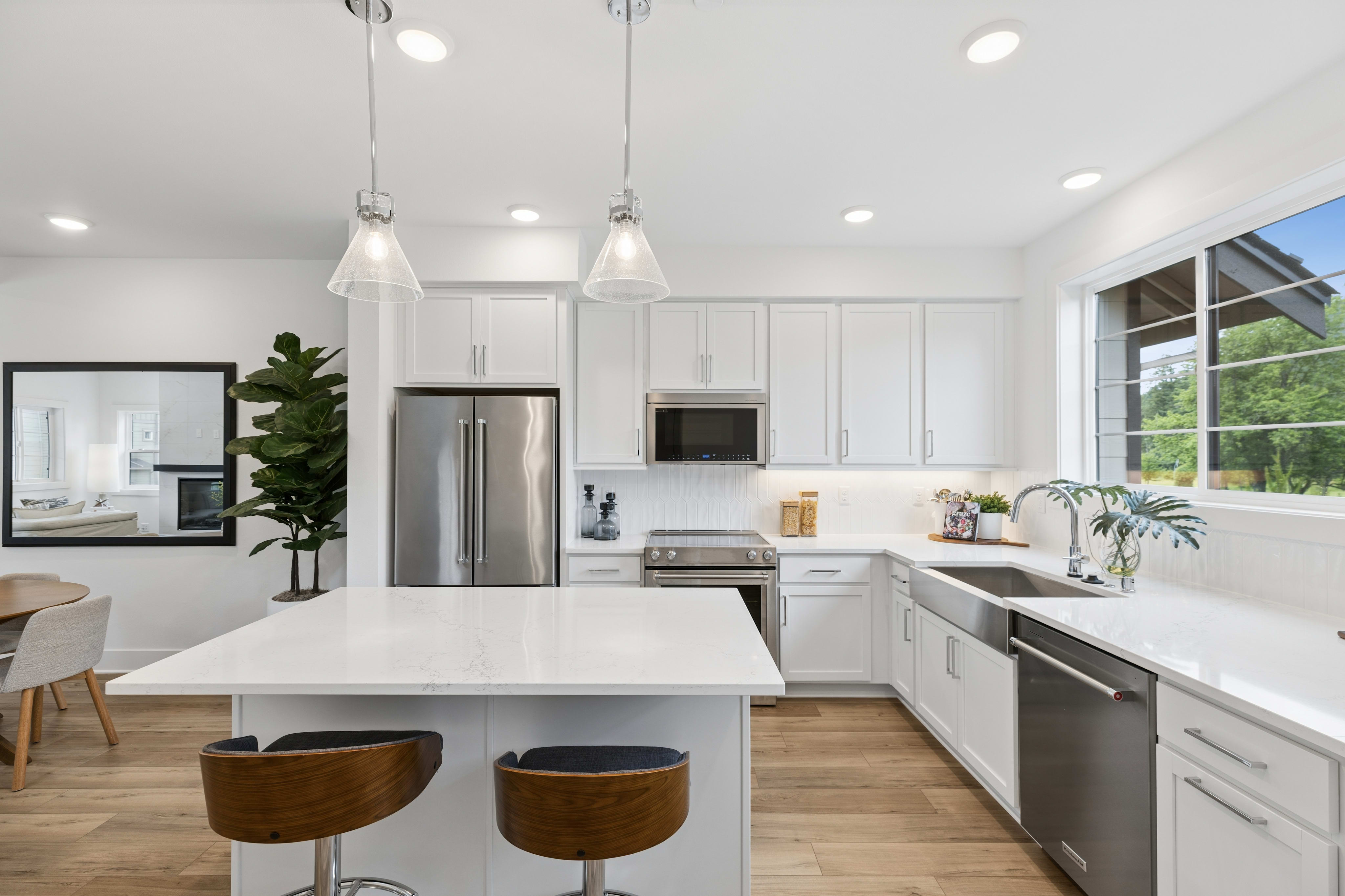 A modern white kitchen with a large island and bar stools in the foreground is complemented by a bright window overlooking a lush green landscape in the background.