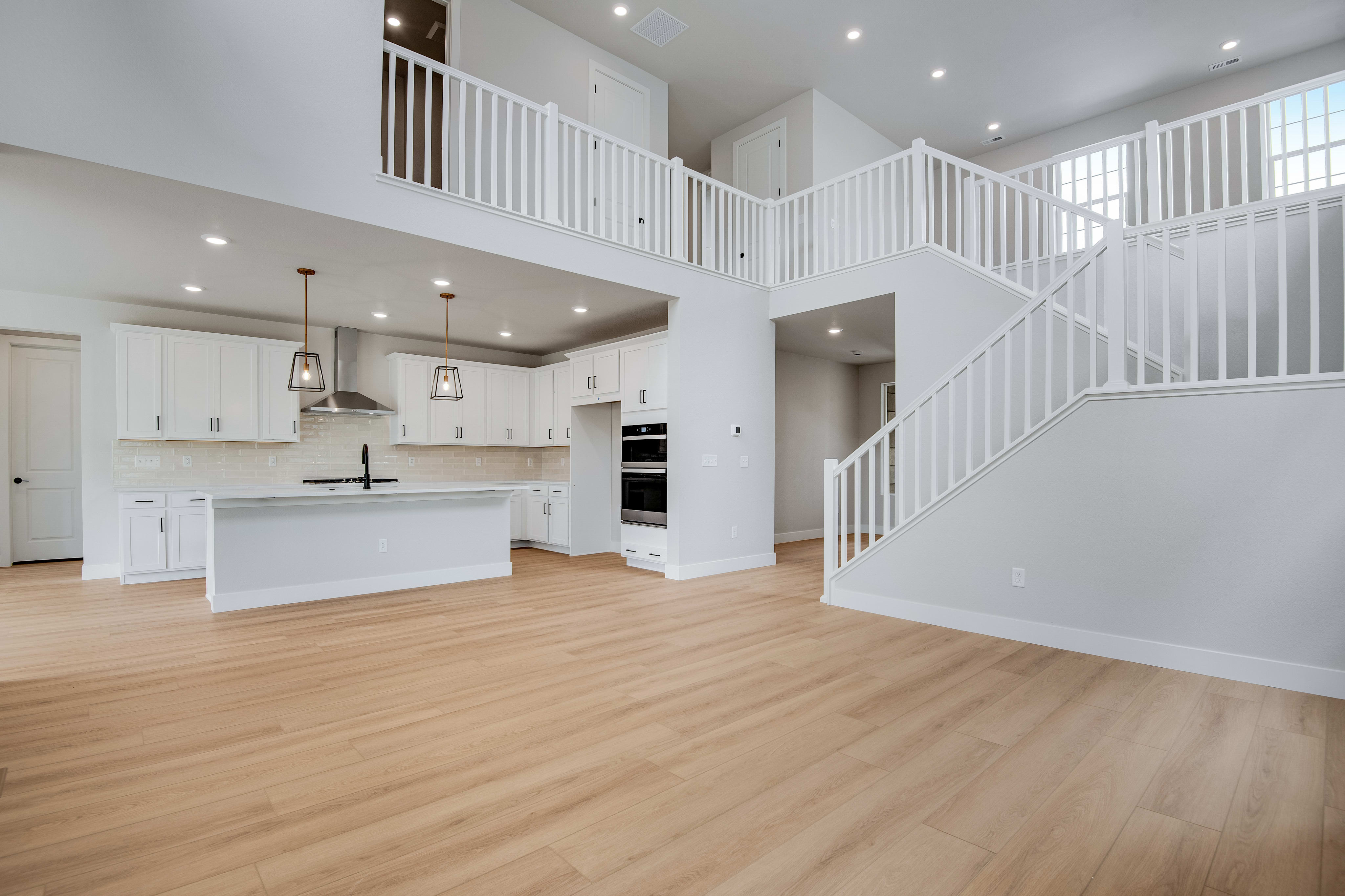 Light wood flooring fills the foreground of a spacious, modern open-concept living area with a white kitchen and a white staircase leading to a loft in the background.