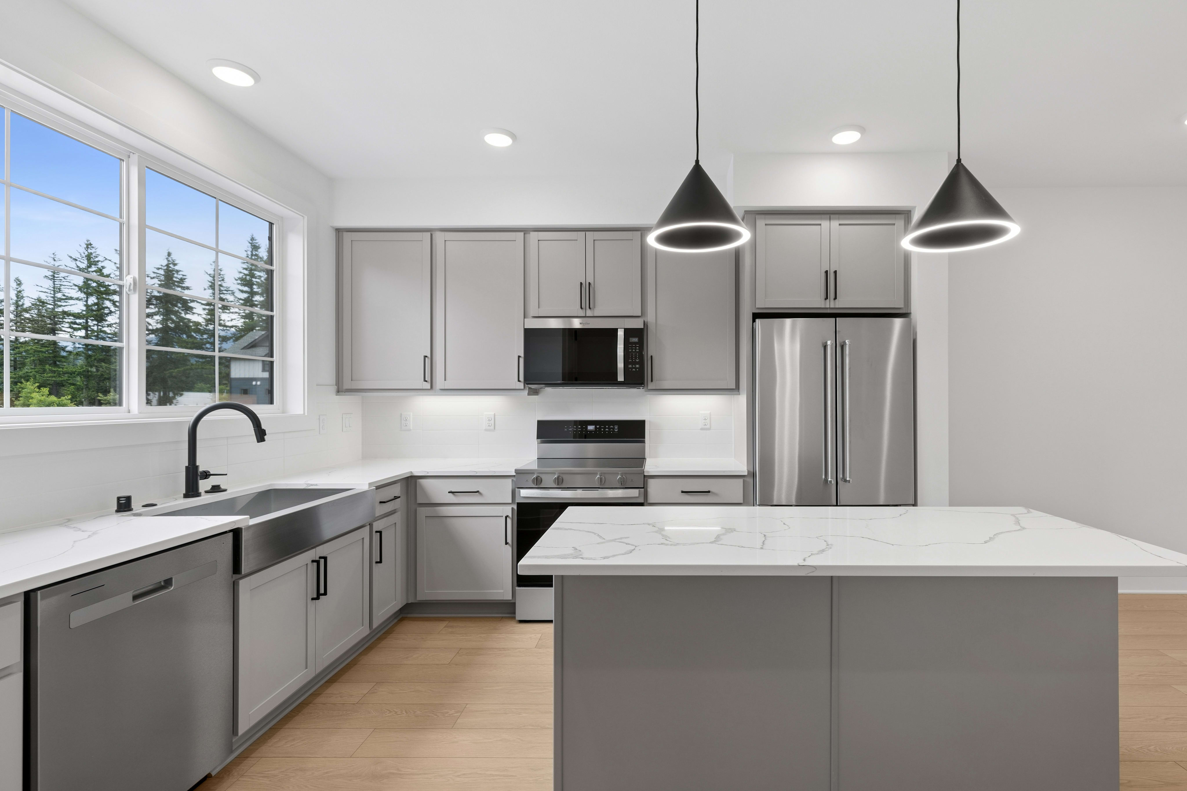 A modern kitchen with a large sink and dishwasher in the foreground and stainless steel appliances and gray cabinets in the background, framed by a large window overlooking trees.
