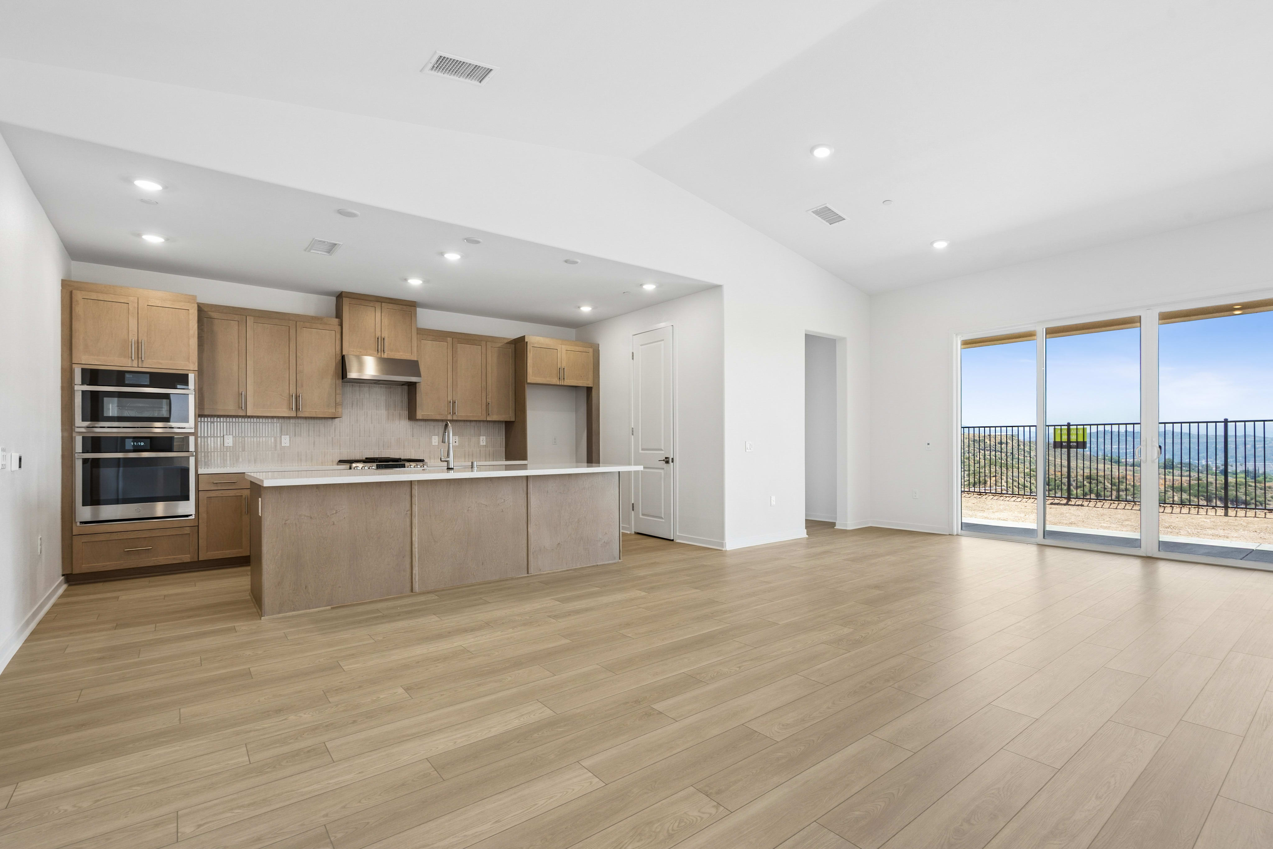 A modern, empty kitchen with light wood cabinets and a large island is in the foreground, while a panoramic view of a scenic landscape is visible through expansive windows in the background.