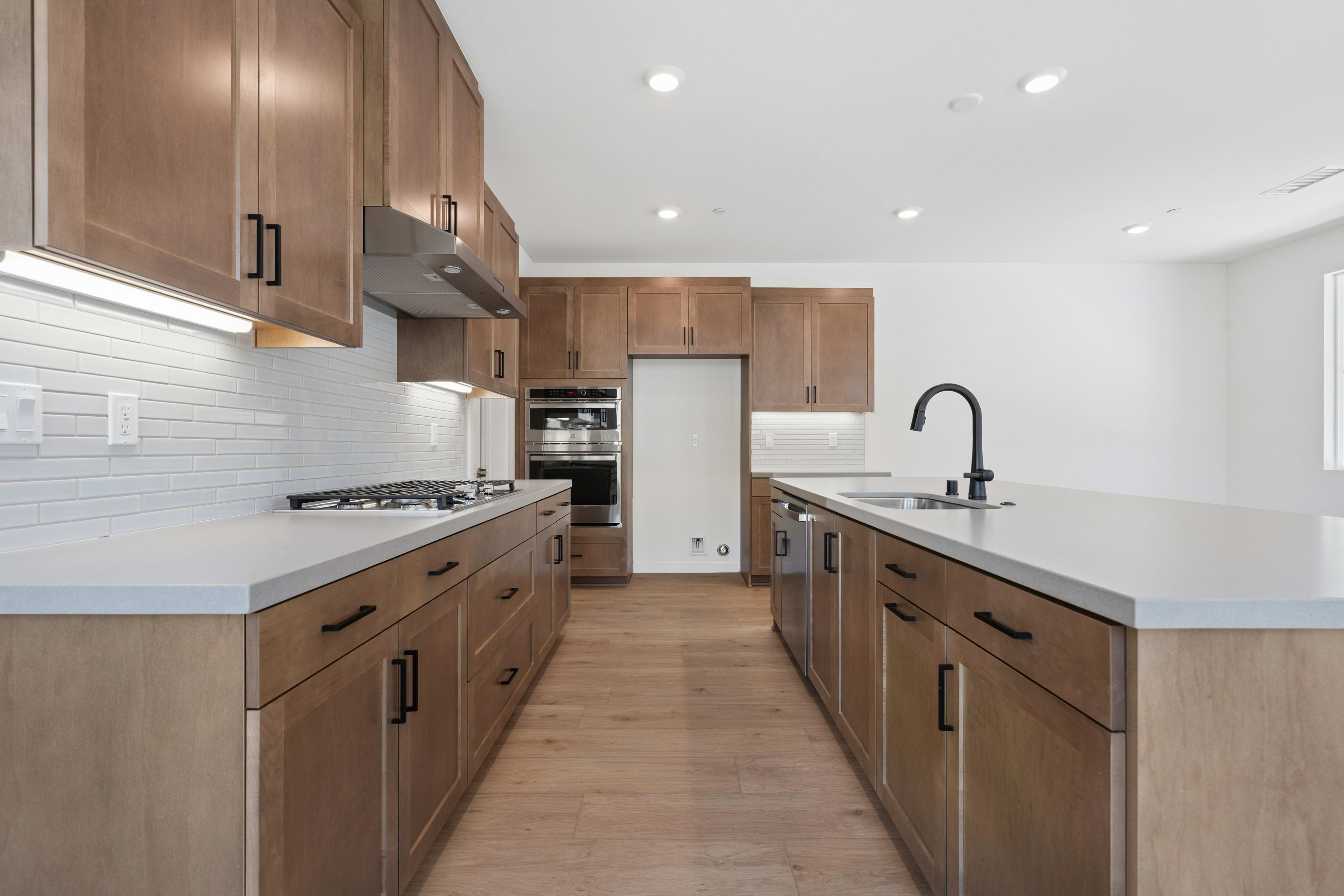 A modern kitchen with light wood cabinets and white countertops features a gas range in the foreground and a sink island in the background, all set against a white-walled room.