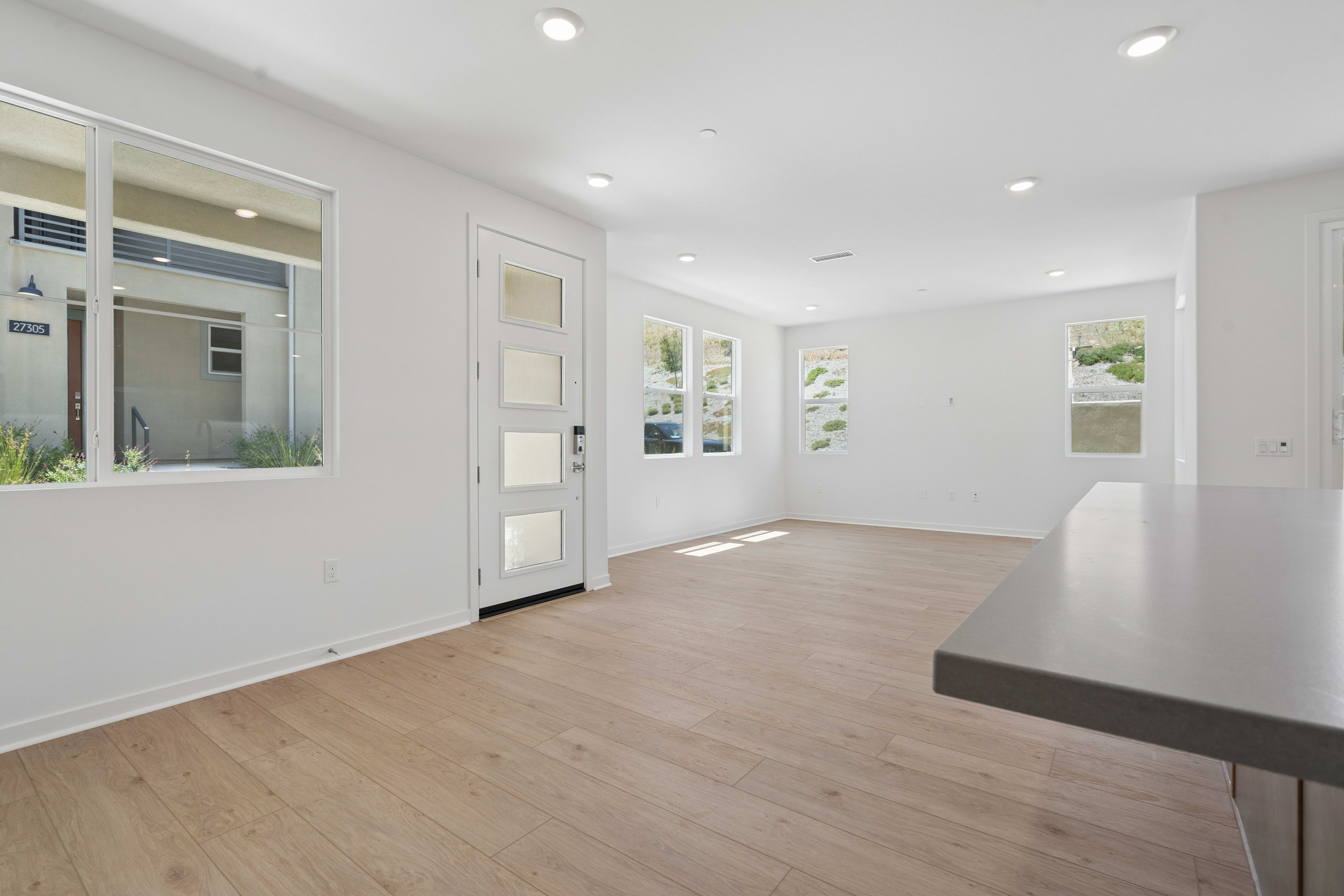 A modern living room with light wood floors in the foreground and large windows revealing a bright, outdoor view in the background.