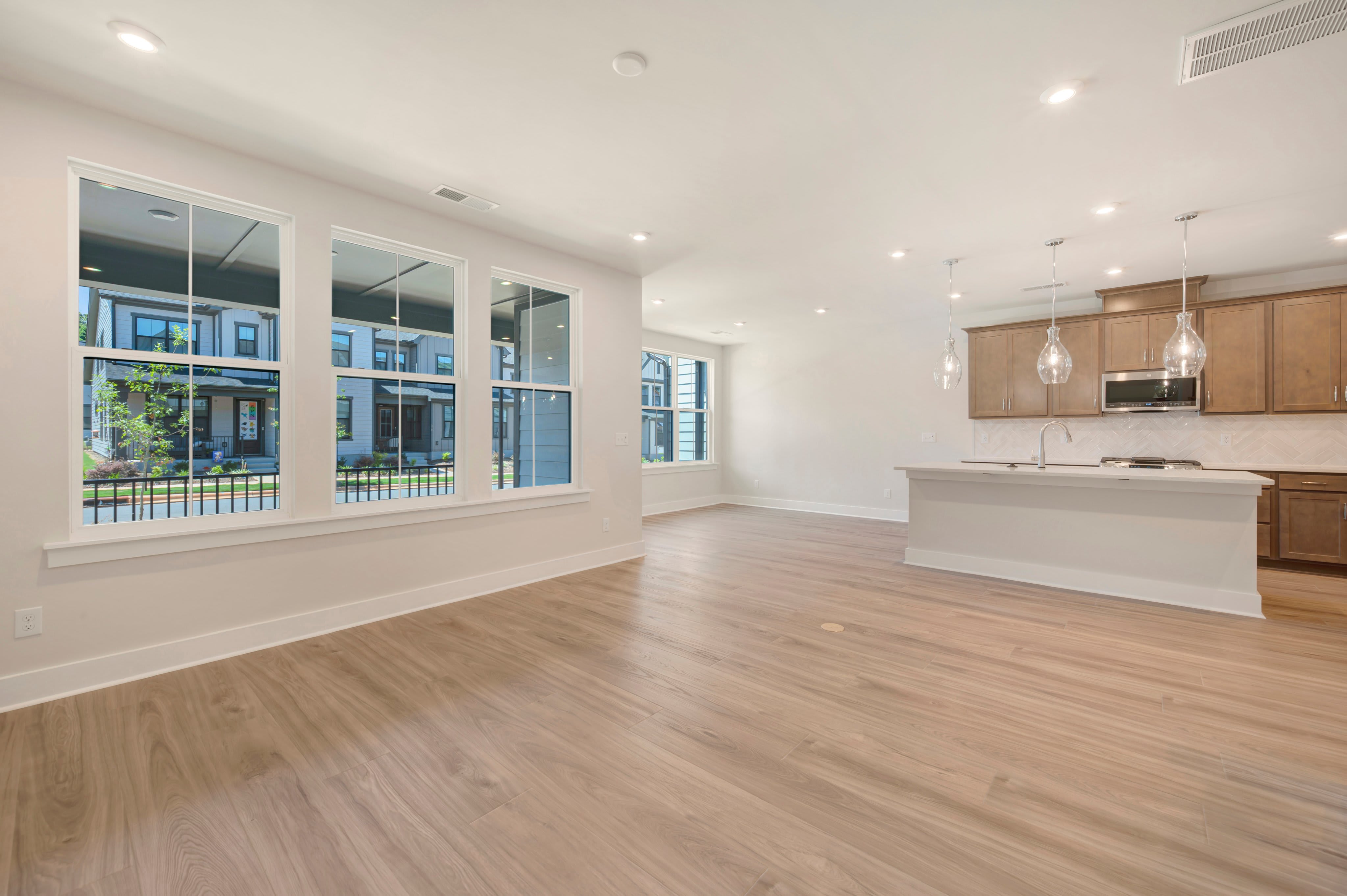 A bright, modern living room with large windows overlooking a street of houses leads into a kitchen with a breakfast bar and pendant lights.