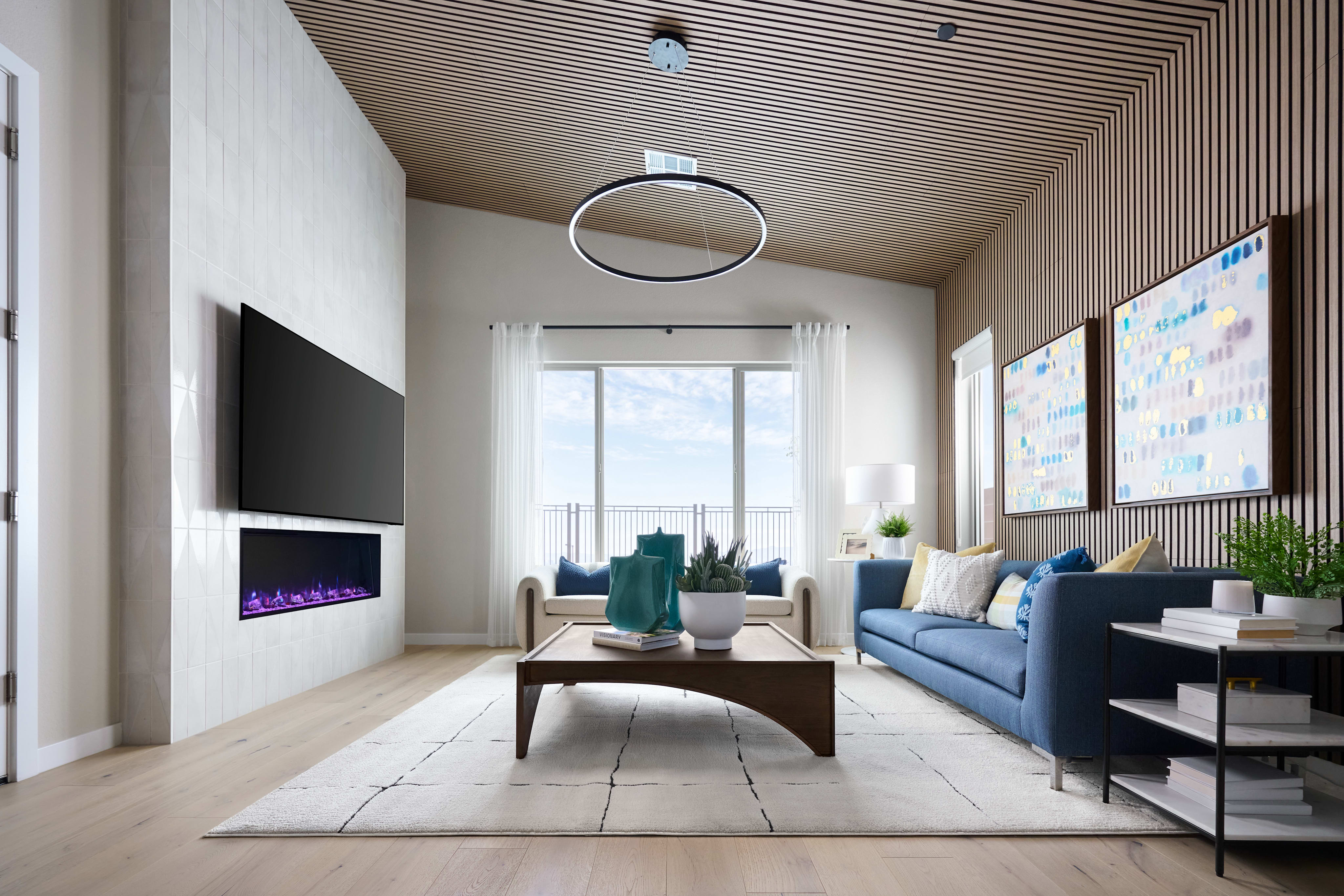 A modern living room with a blue sofa and coffee table in the foreground, and a large window with a view of the sky in the background.