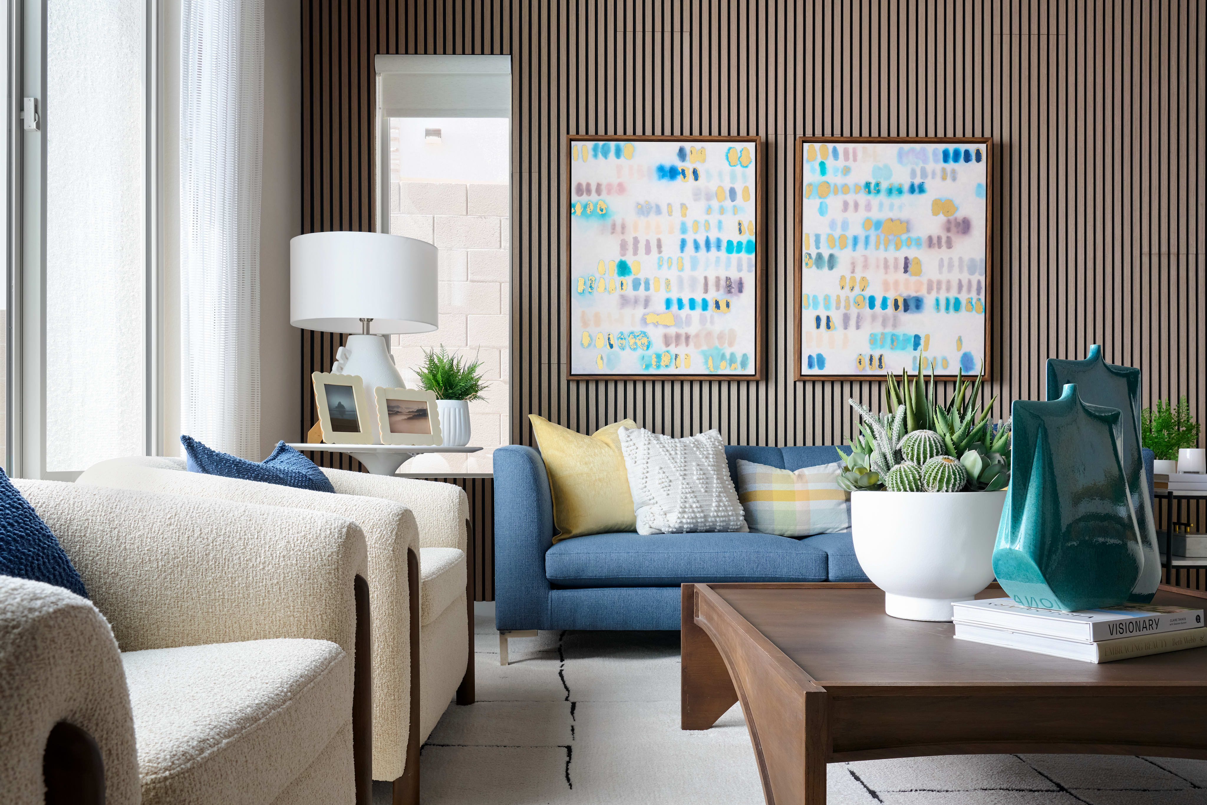Two plush armchairs and a blue sofa with colorful pillows sit in the foreground, while the background features two abstract paintings on a textured wall.