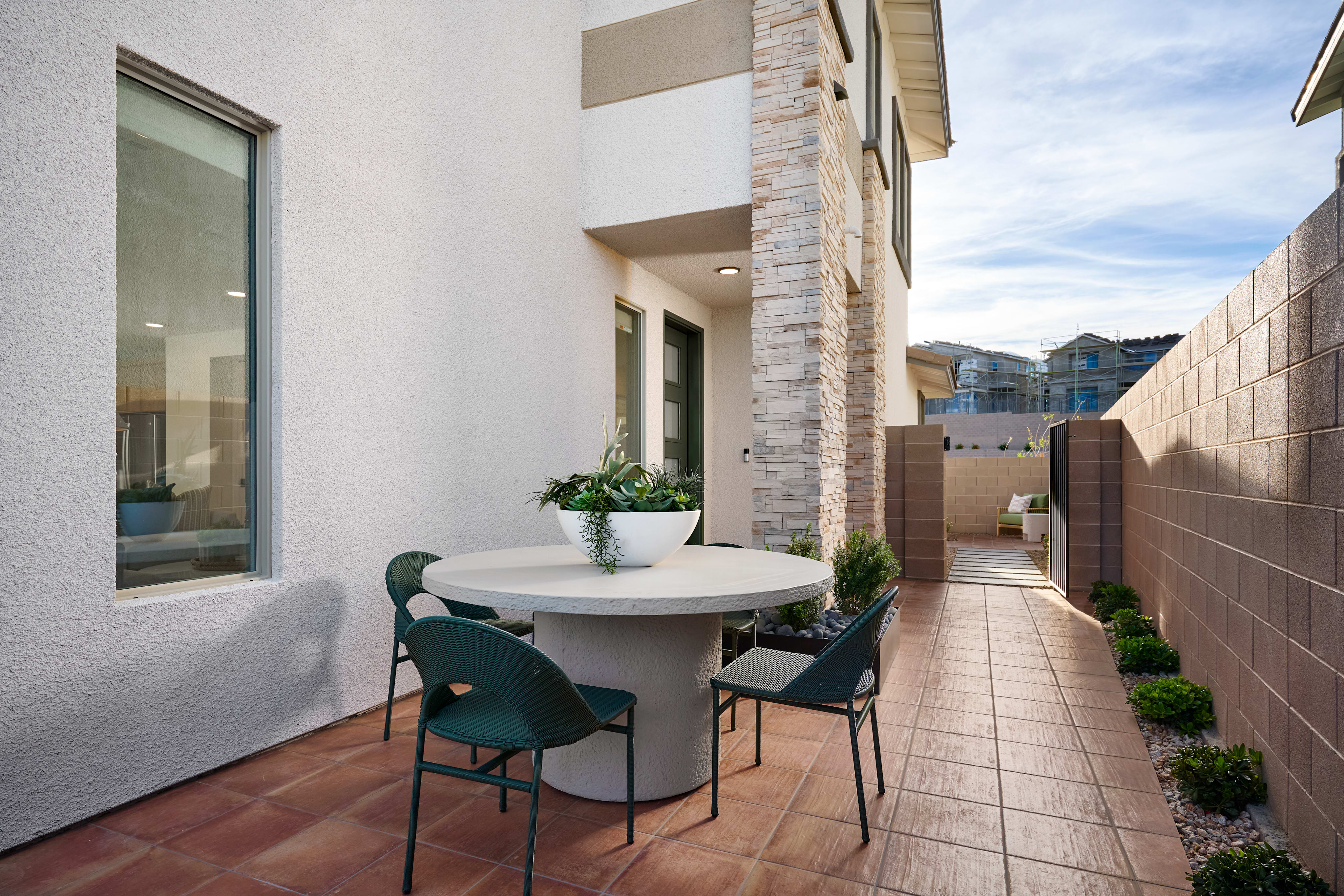 A round table with four chairs sits on a tiled patio in the foreground, while a narrow walkway leads to houses in the background.
