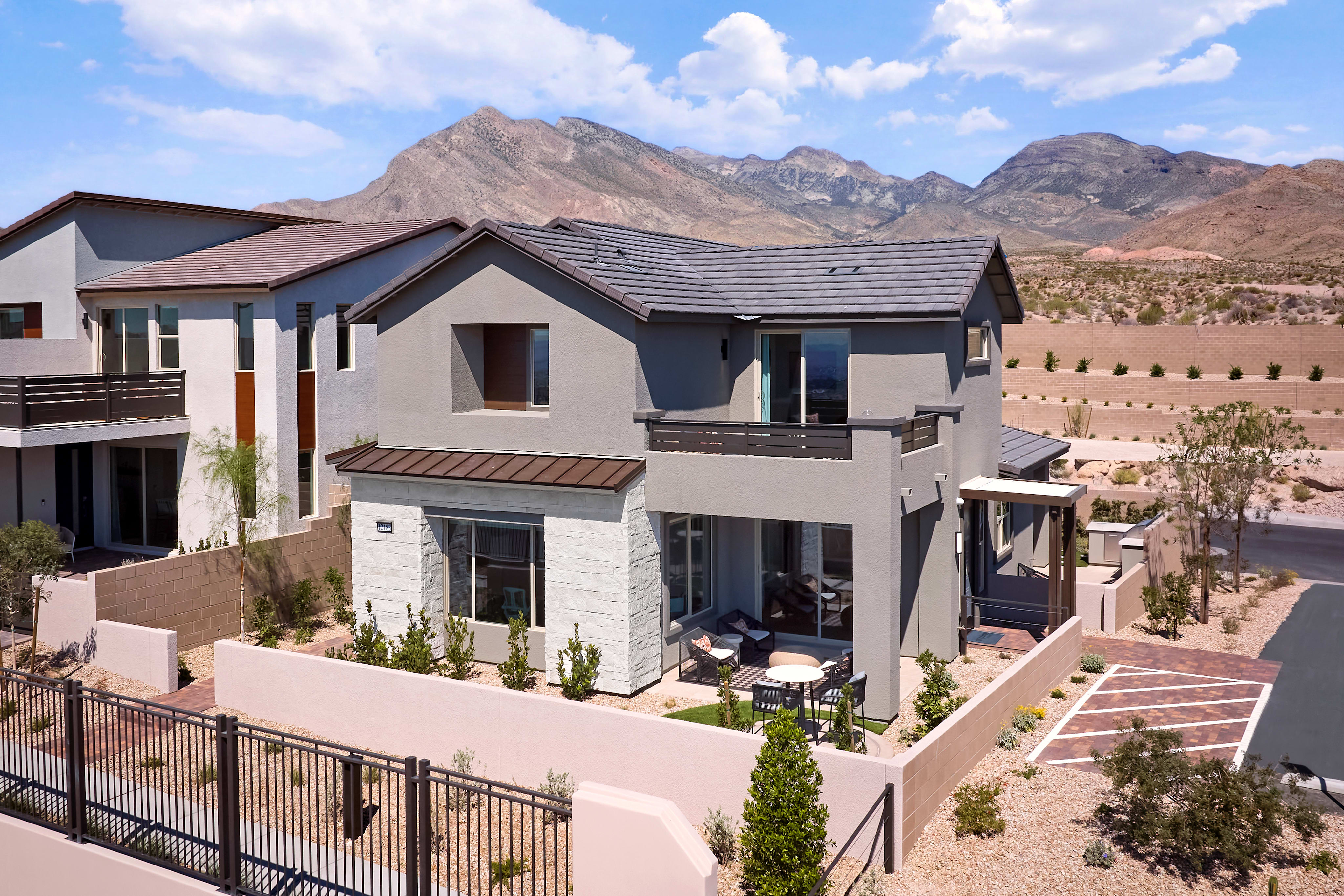 A modern gray house with a small patio sits in the foreground, while rugged, arid mountains rise in the background under a bright blue sky.
