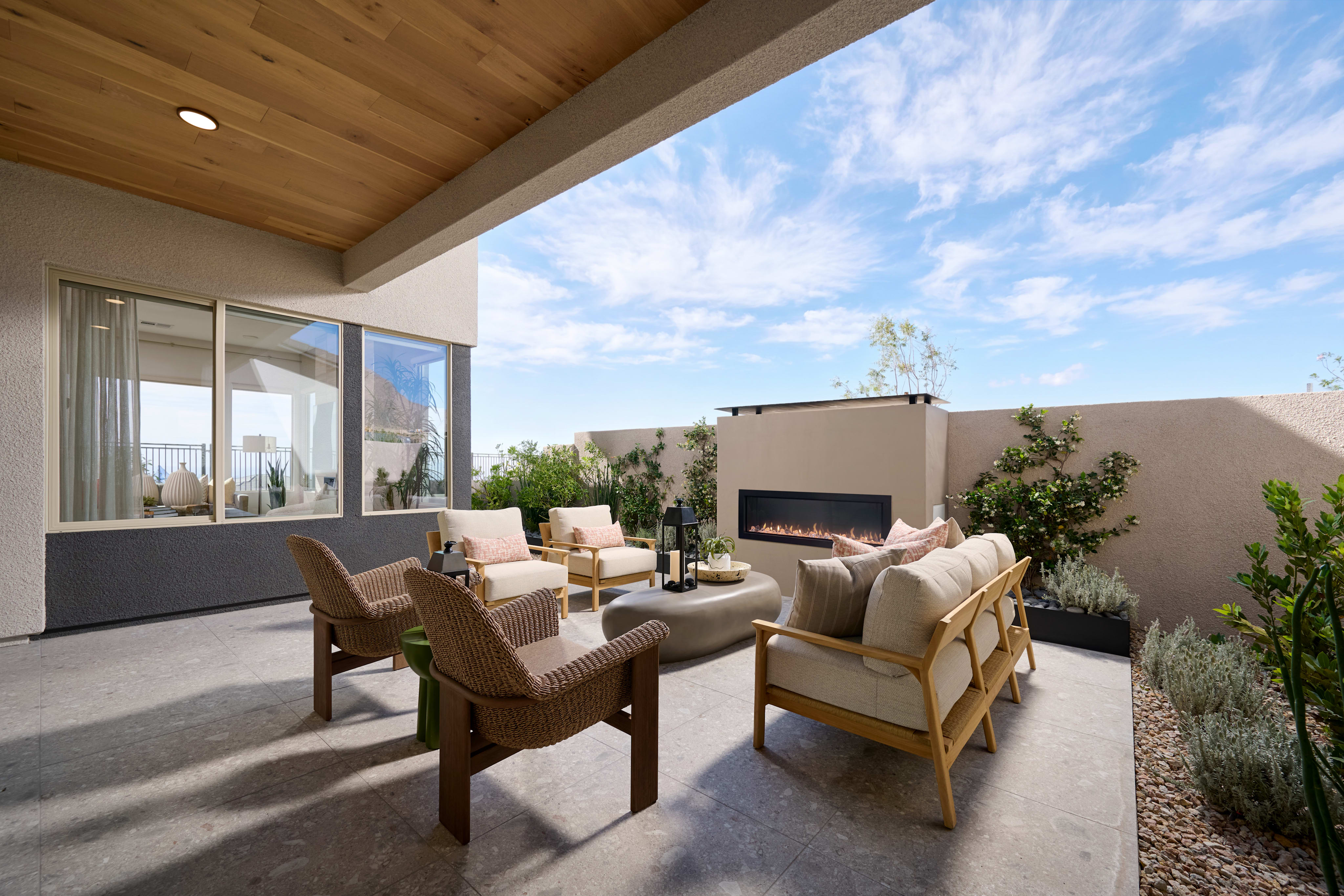 A modern patio with comfortable seating and a fireplace is set against a backdrop of a clear blue sky with scattered clouds.