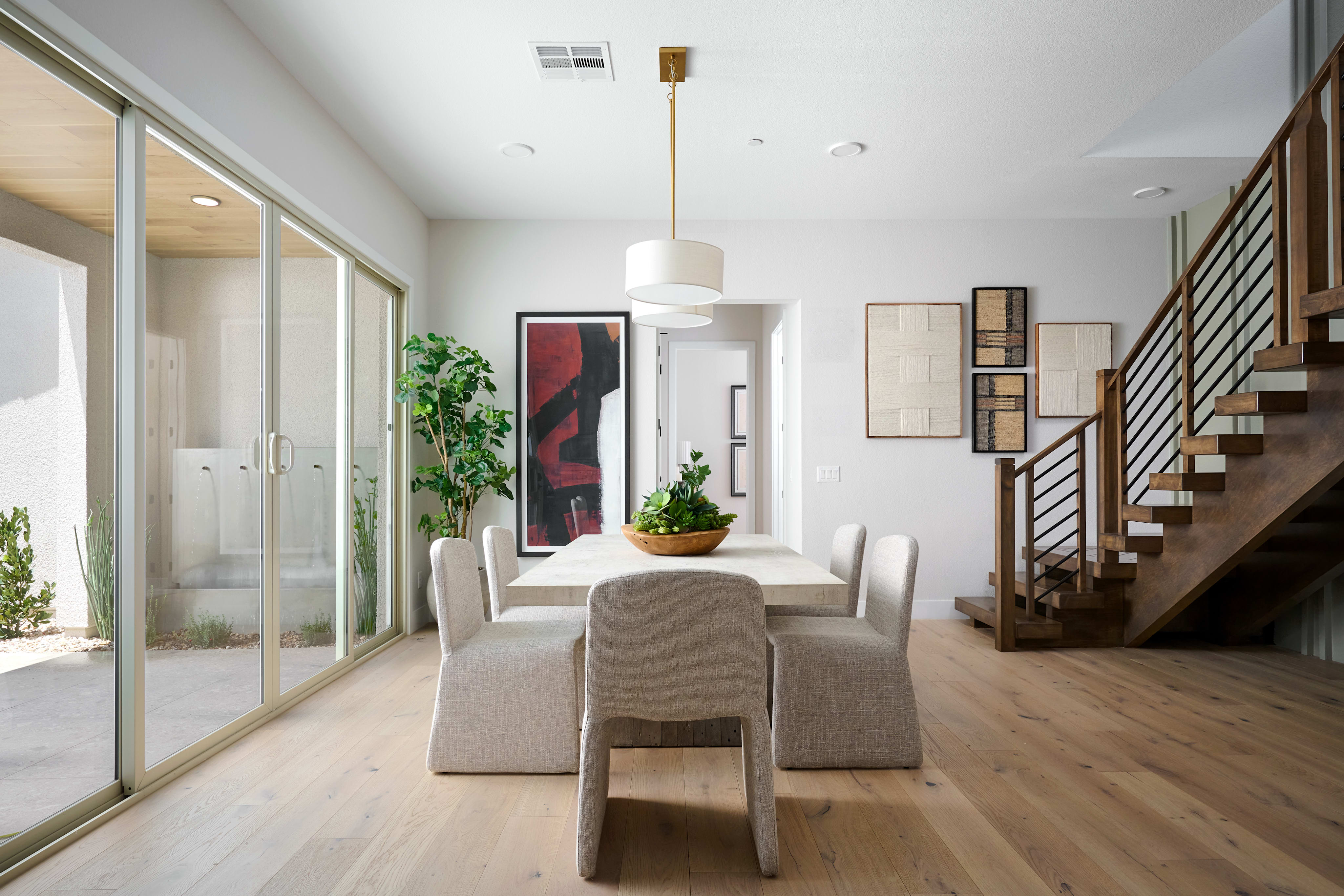 A modern dining room with a light wood floor and a large window overlooking a patio in the foreground, and a wooden staircase and abstract art on the wall in the background.