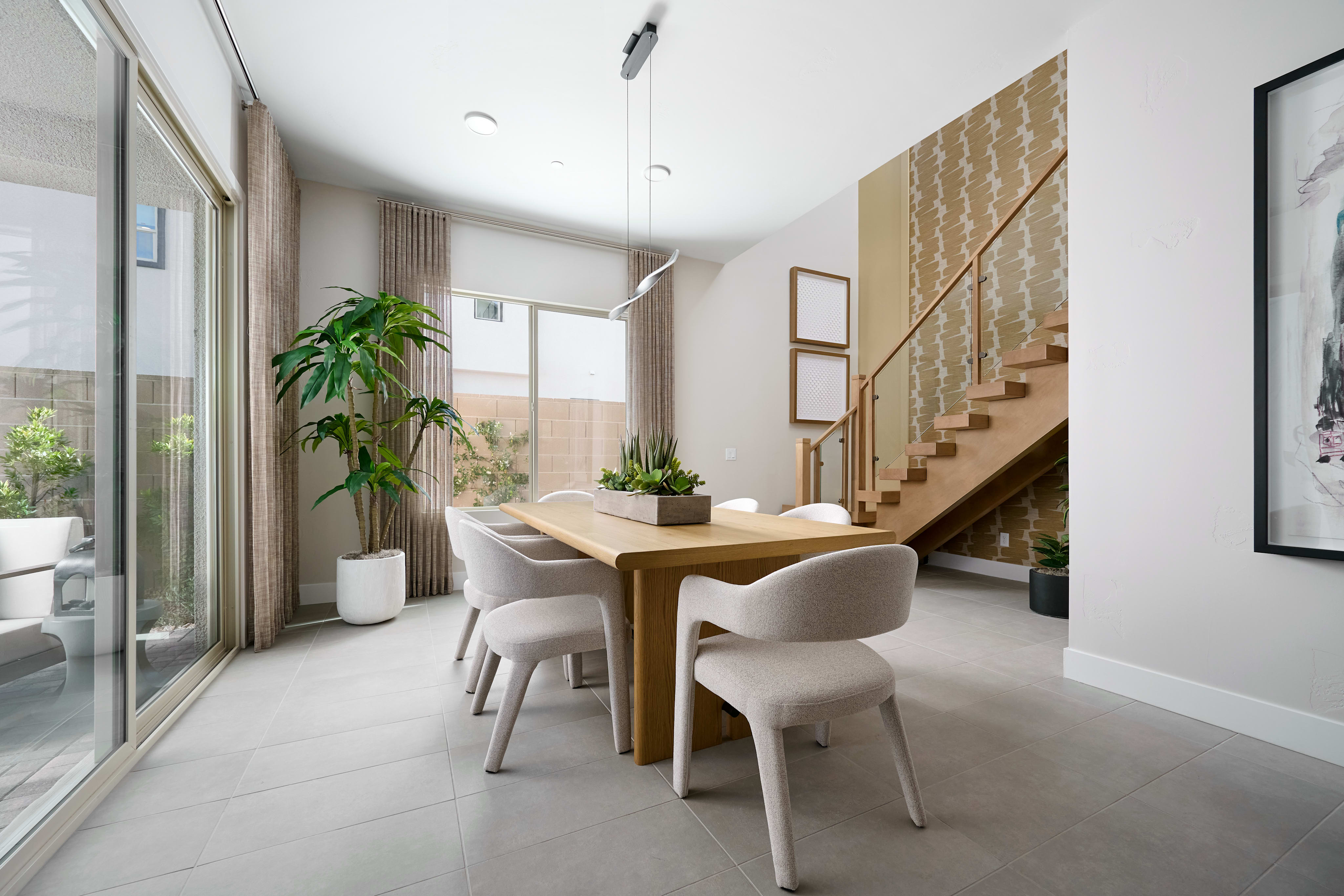 A modern dining room with a light wood table and chairs in the foreground, and a staircase with patterned wallpaper in the background.