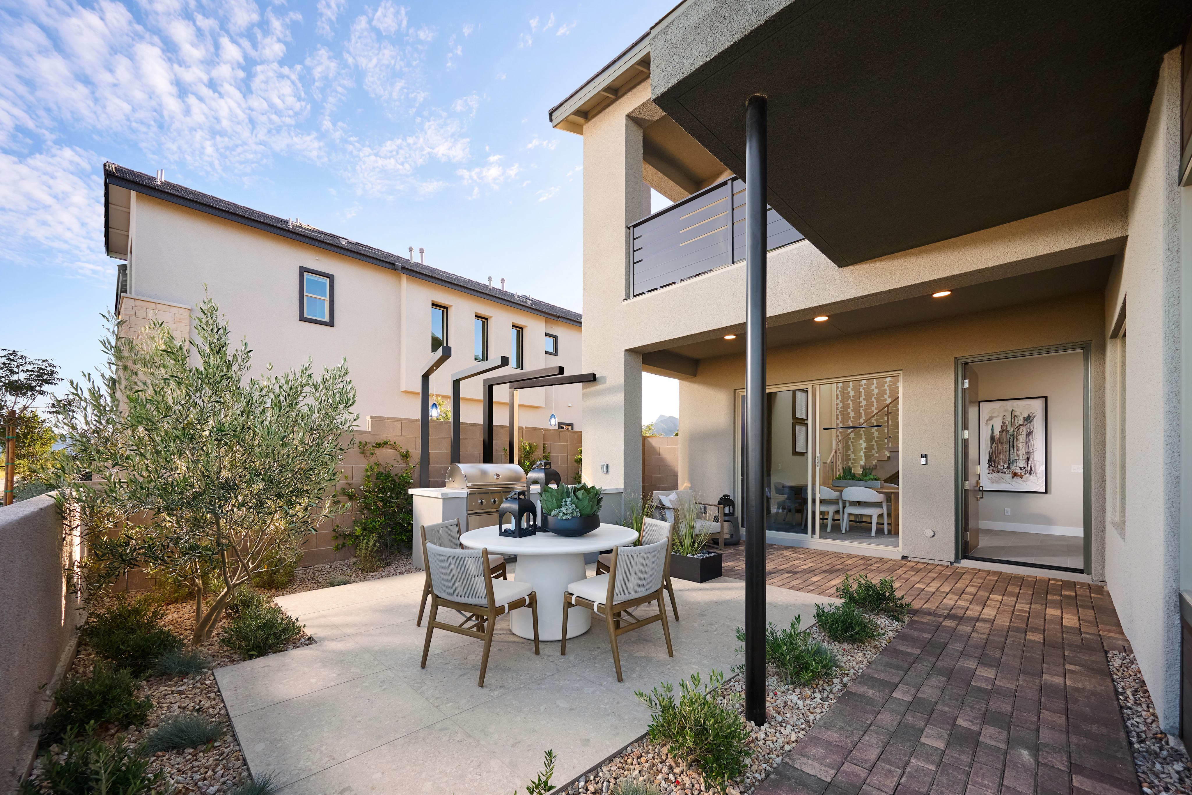 A patio with a dining set and outdoor kitchen is in the foreground, while a modern house and a clear sky are in the background.
