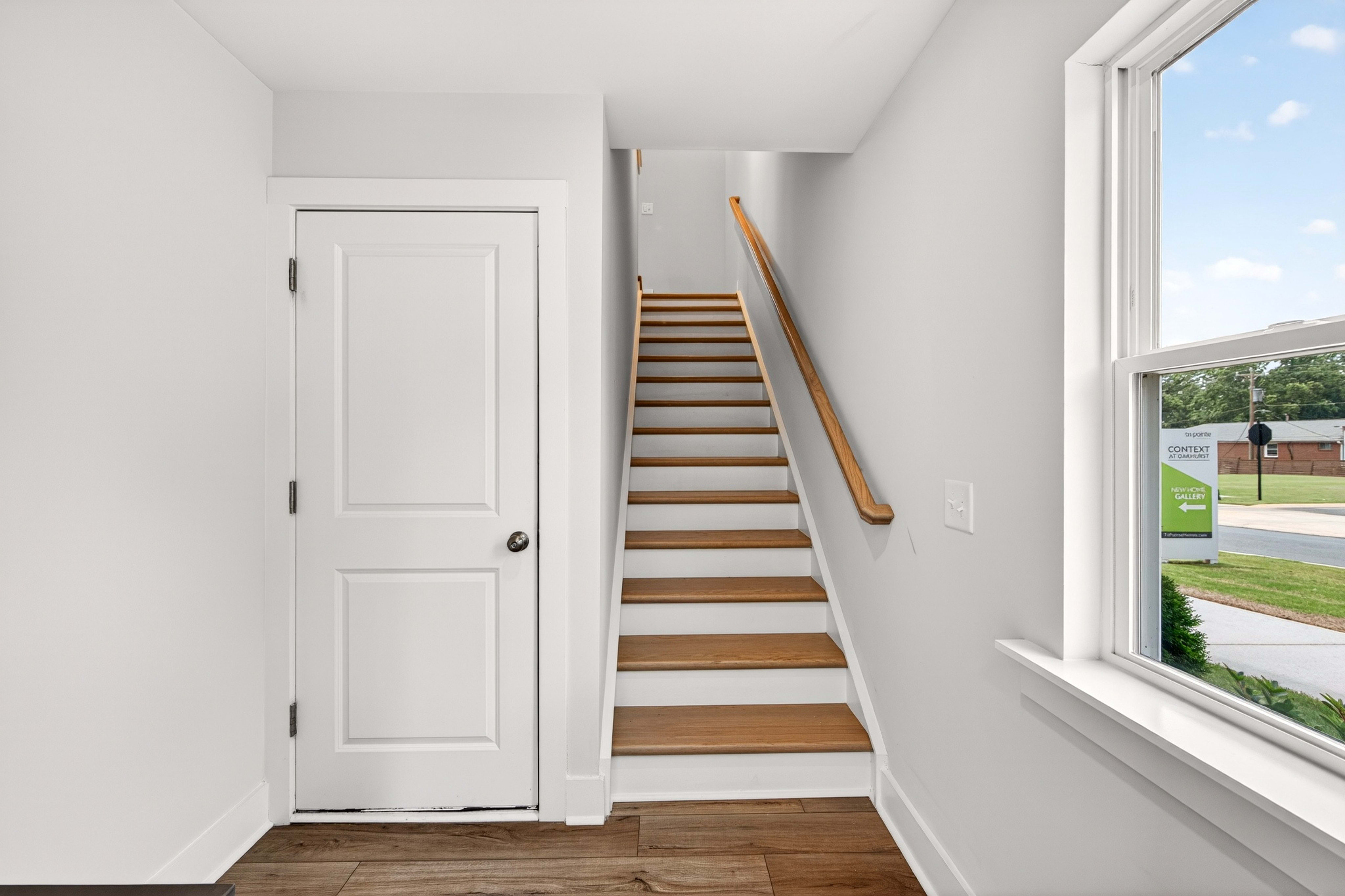 A white door and wooden staircase are in the foreground, while a window reveals a glimpse of the outdoors with a sign and trees in the background.