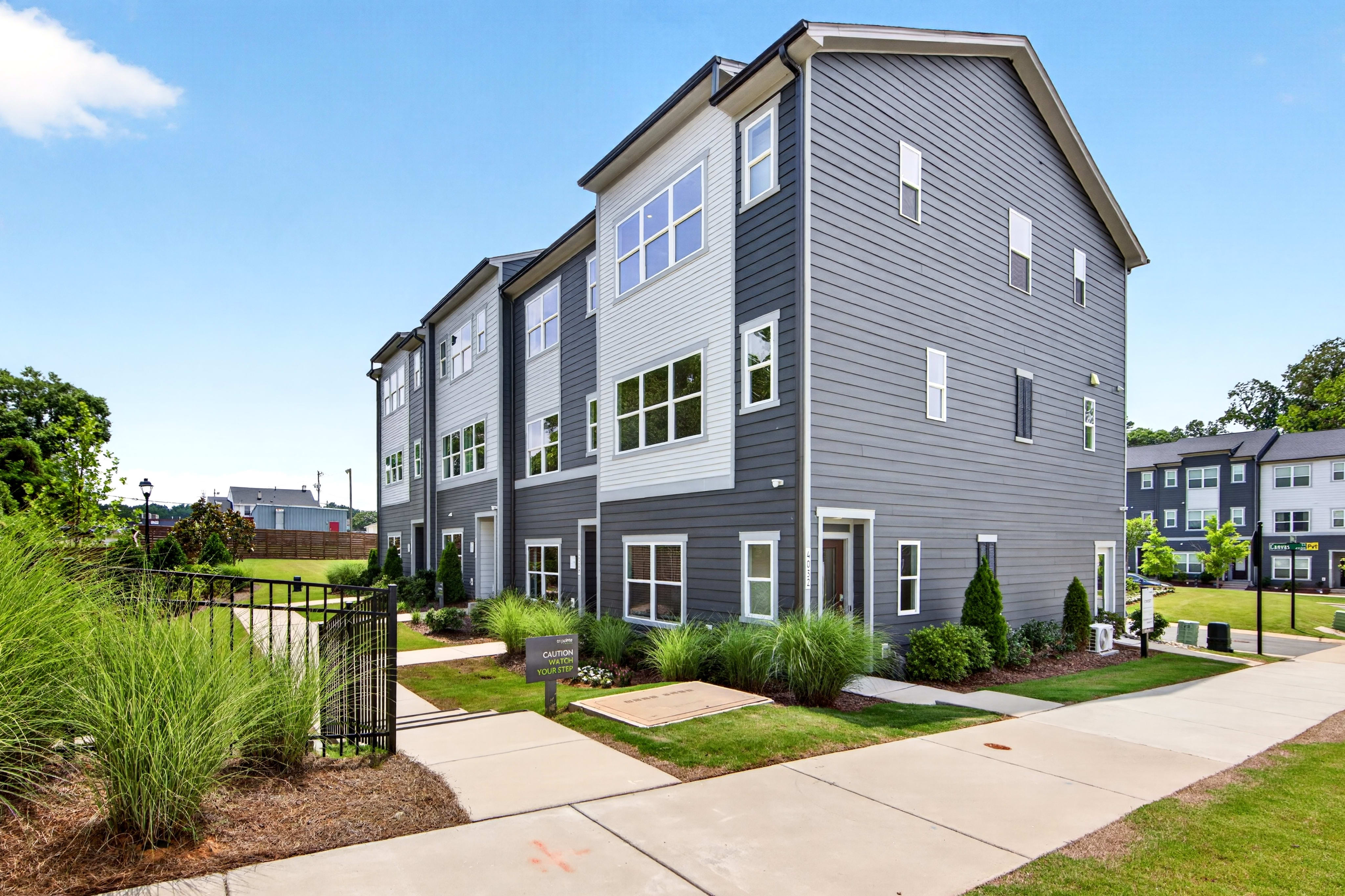 A row of modern townhouses with gray siding and white accents stands under a bright blue sky, with lush green grass and landscaping in the foreground.