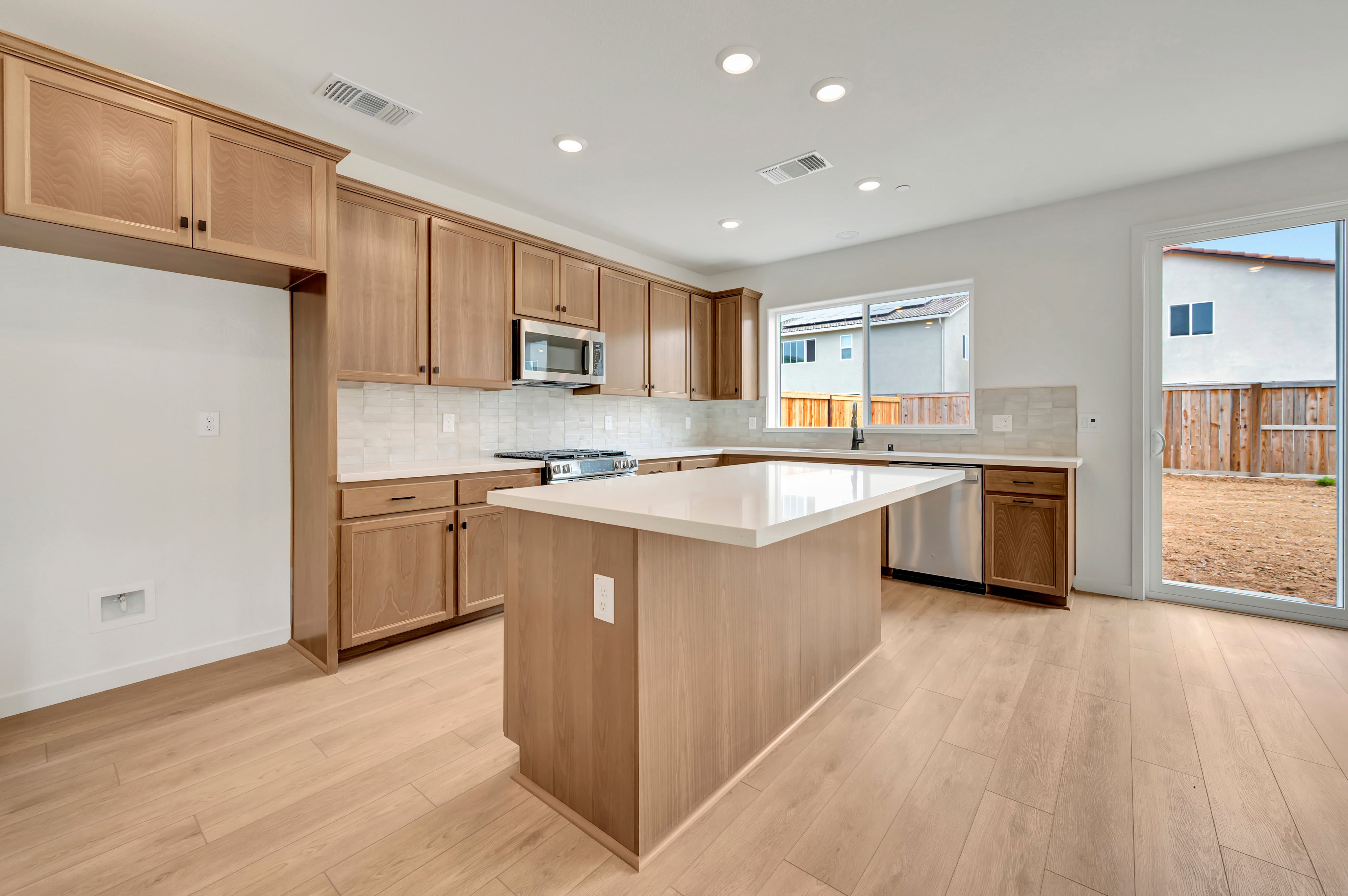 A modern kitchen with a light wood island in the foreground and a backyard visible through a sliding glass door in the background.