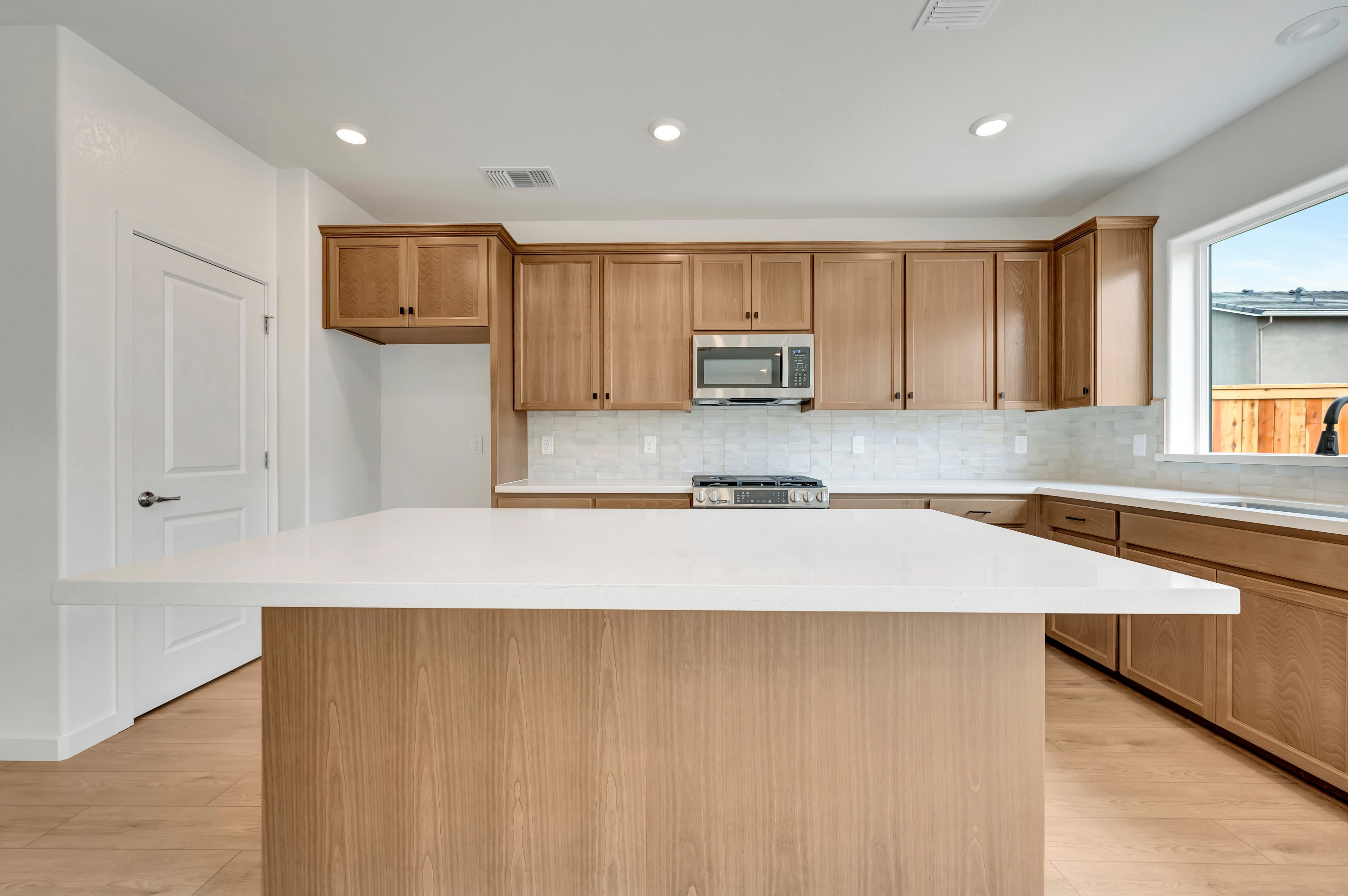 A modern kitchen with a large white island in the foreground and light wood cabinets and a window in the background.