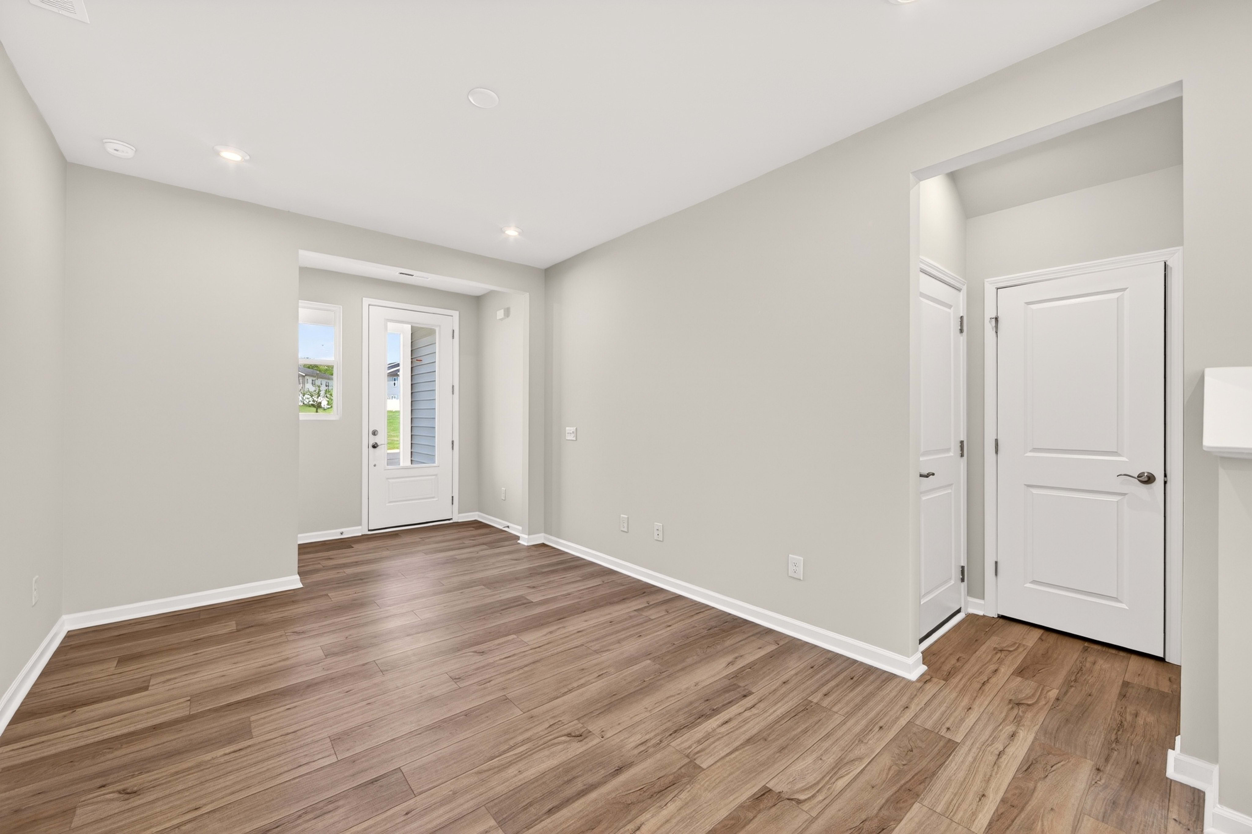 A spacious, empty living room with light gray walls and wood-look flooring in the foreground, and a white front door and interior doors in the background.