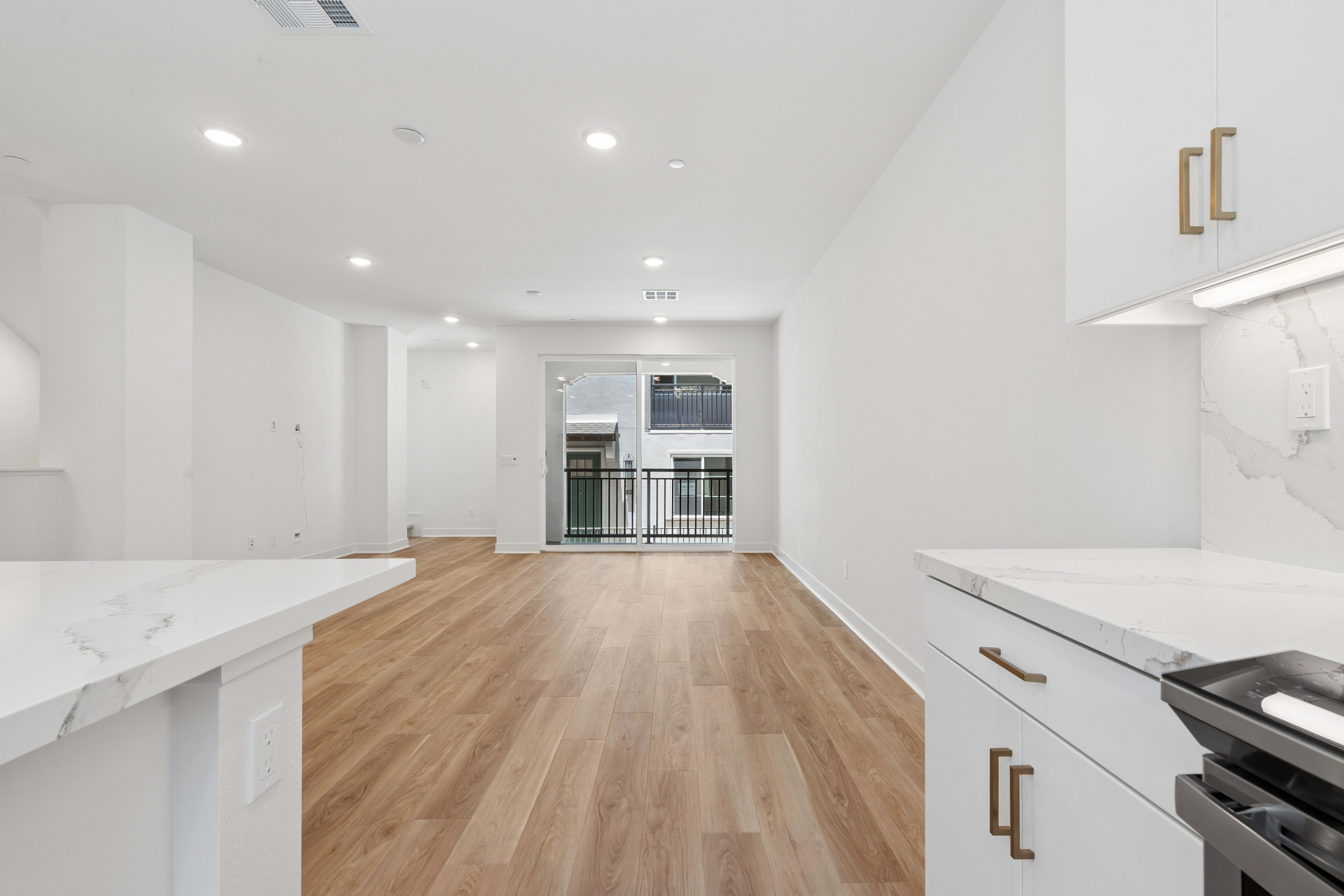 A modern, empty living space with light wood floors and white walls is visible, with a kitchen counter in the foreground and a balcony with a railing in the background.