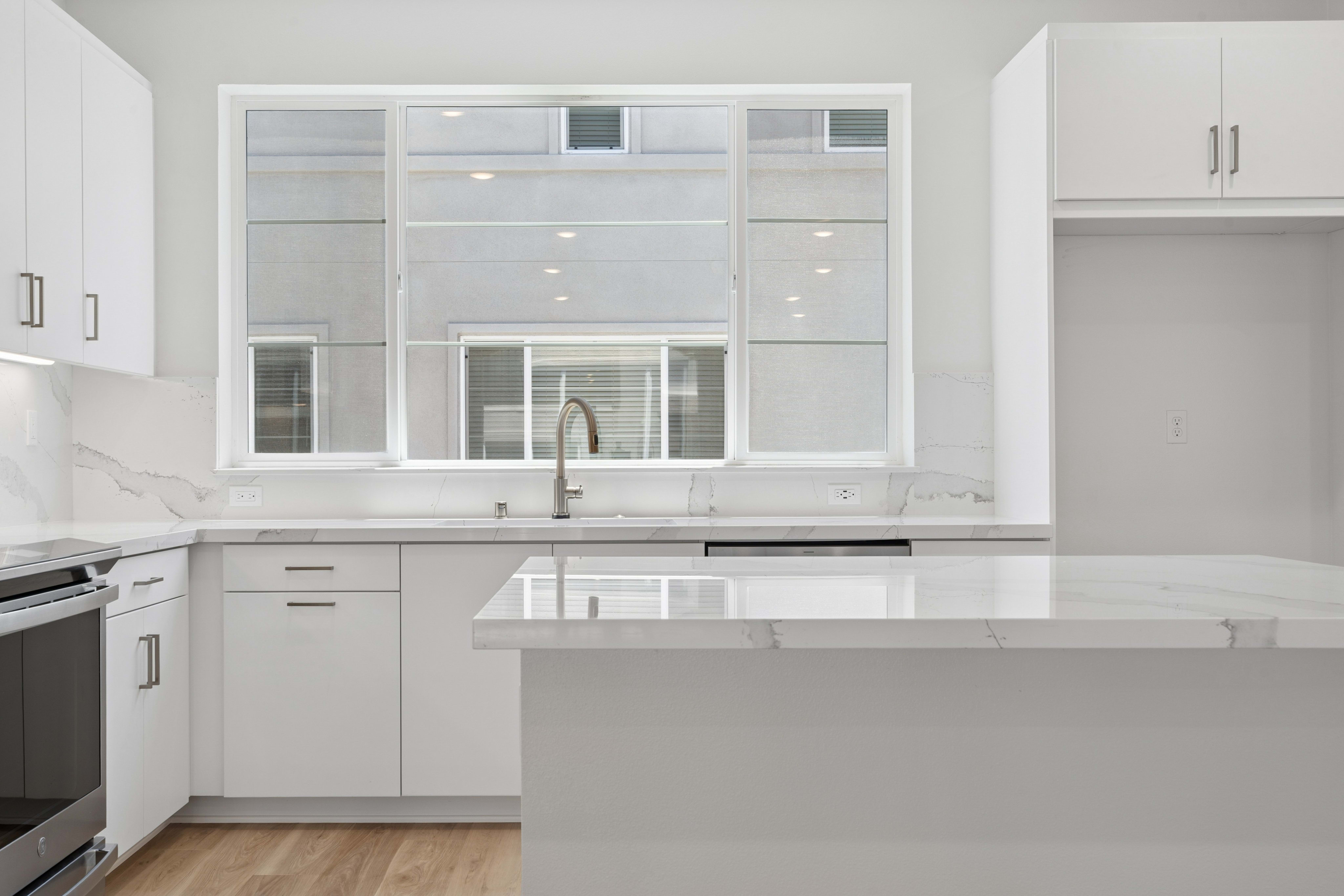 A modern white kitchen with a marble island in the foreground and a large window reflecting the exterior in the background.
