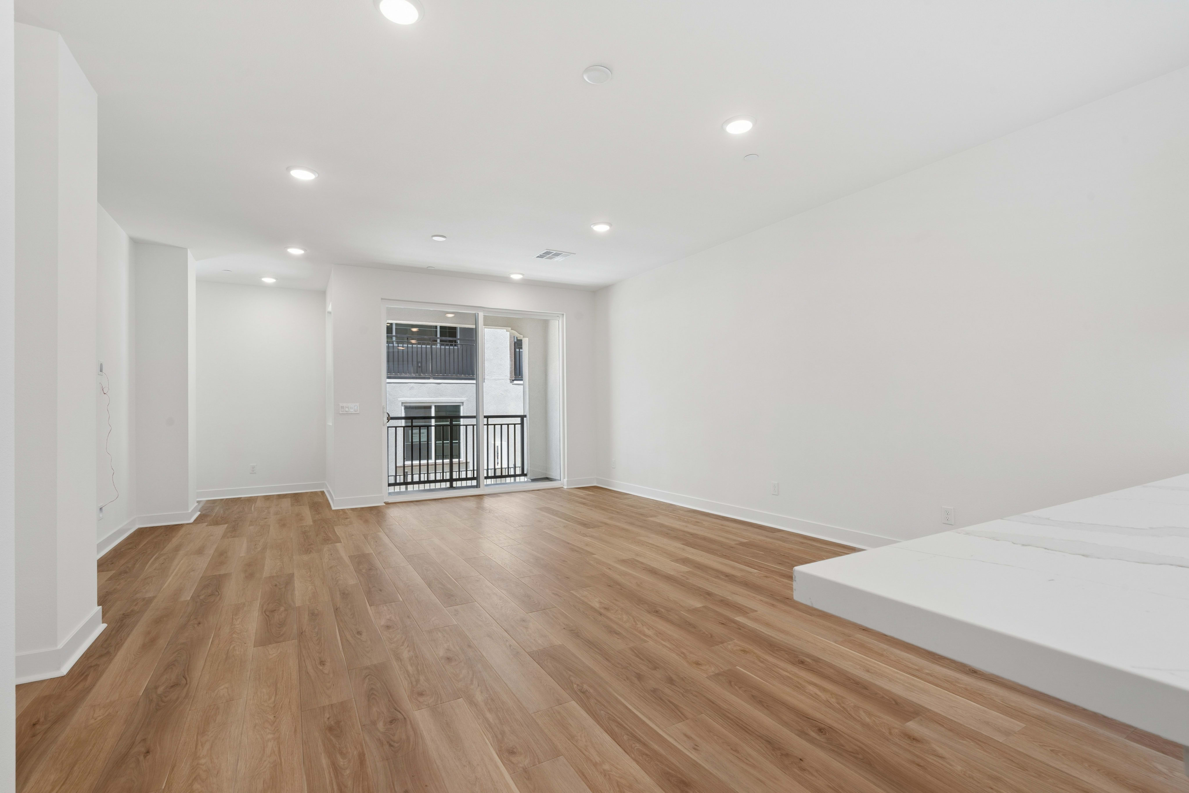 A bright, empty living room with light wood floors in the foreground and a sliding glass door leading to a balcony in the background.